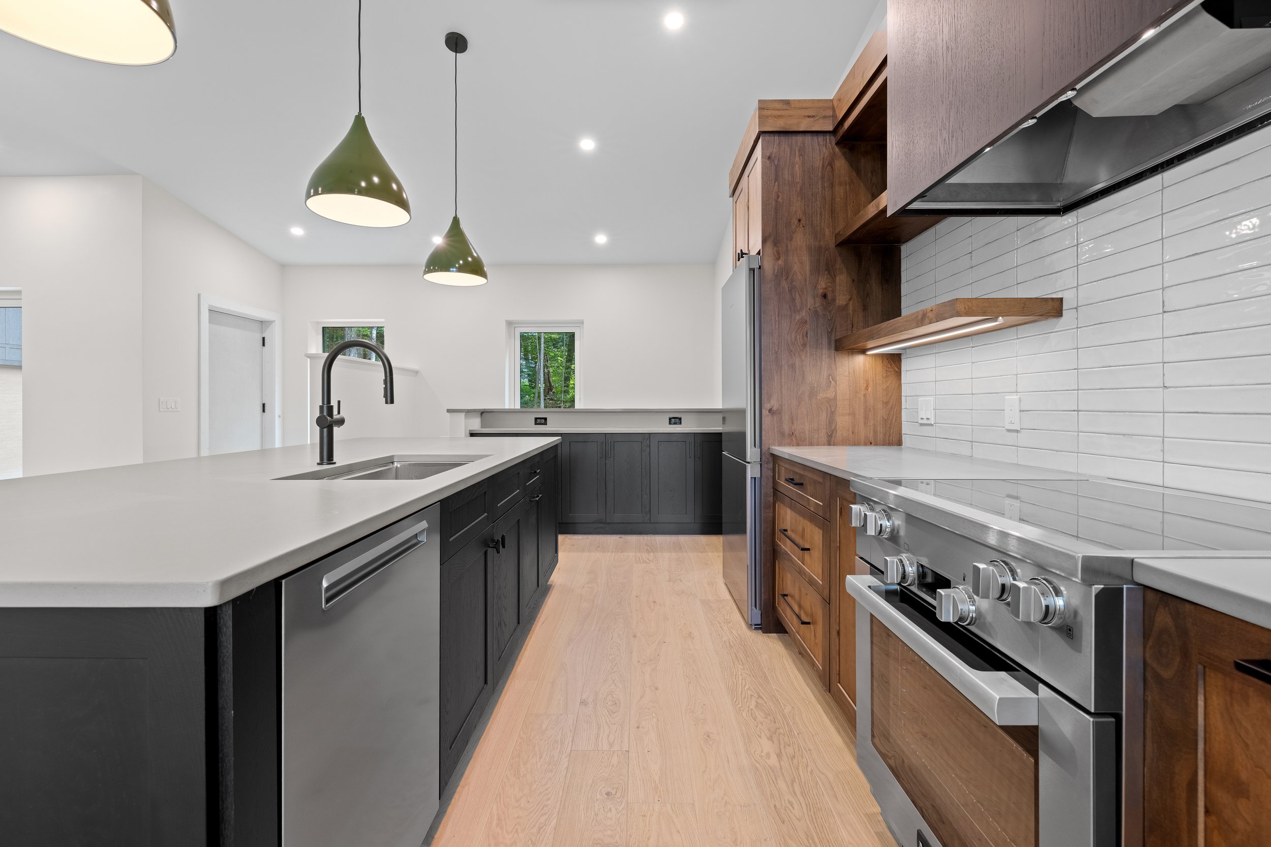 Modern kitchen with white countertops, dark island cabinetry, wood cabinets, subway tile backsplash, stainless steel appliances, and green pendant lights