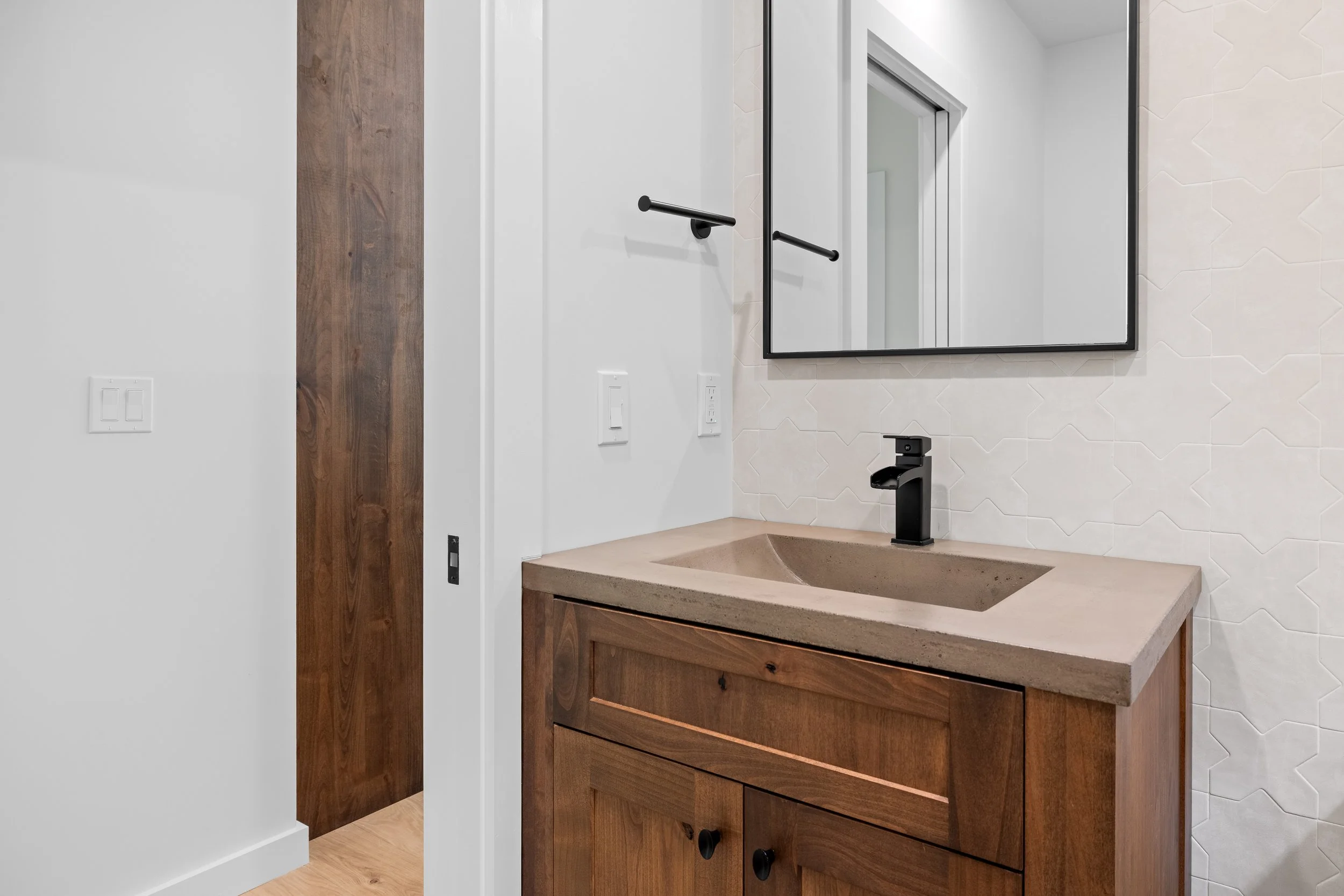 Modern bathroom vanity with wood cabinetry, concrete countertop, black faucet, geometric white tile backsplash, and framed mirror