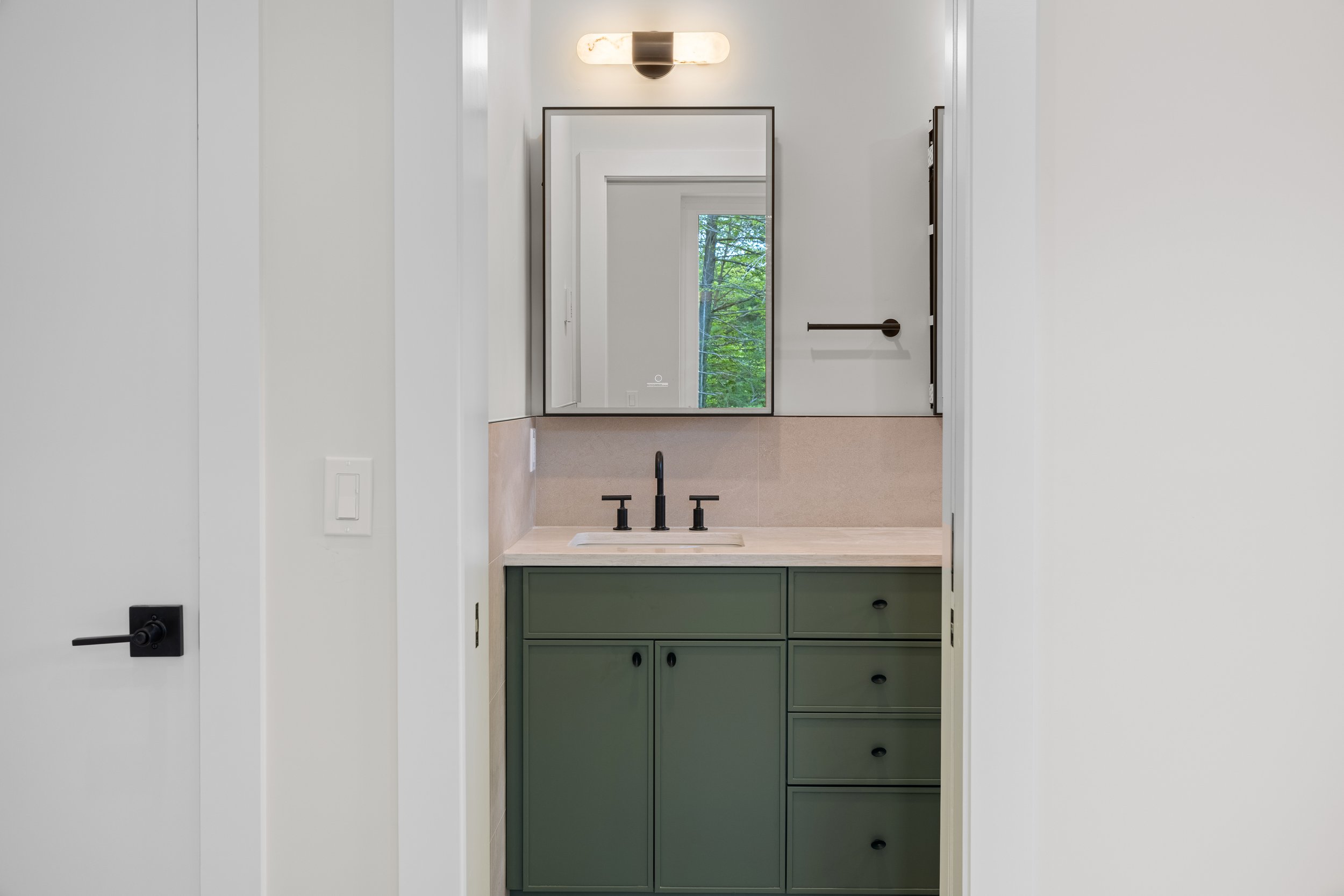Modern bathroom vanity with dark green cabinets, marble countertop, black fixtures, wall mirror, and window overlooking trees