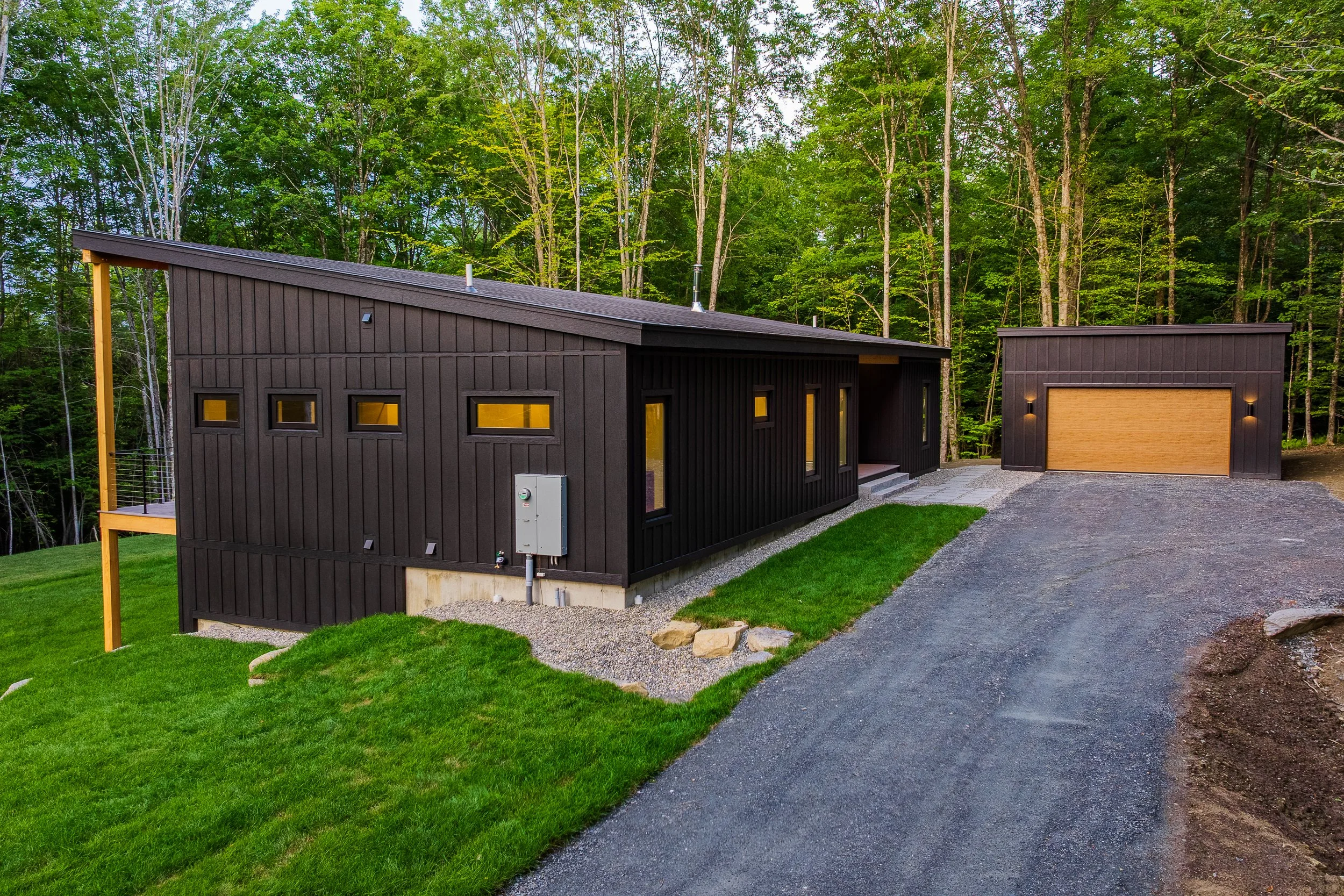 Modern dark-sided house with attached garage, gravel driveway, and manicured lawn in a wooded setting