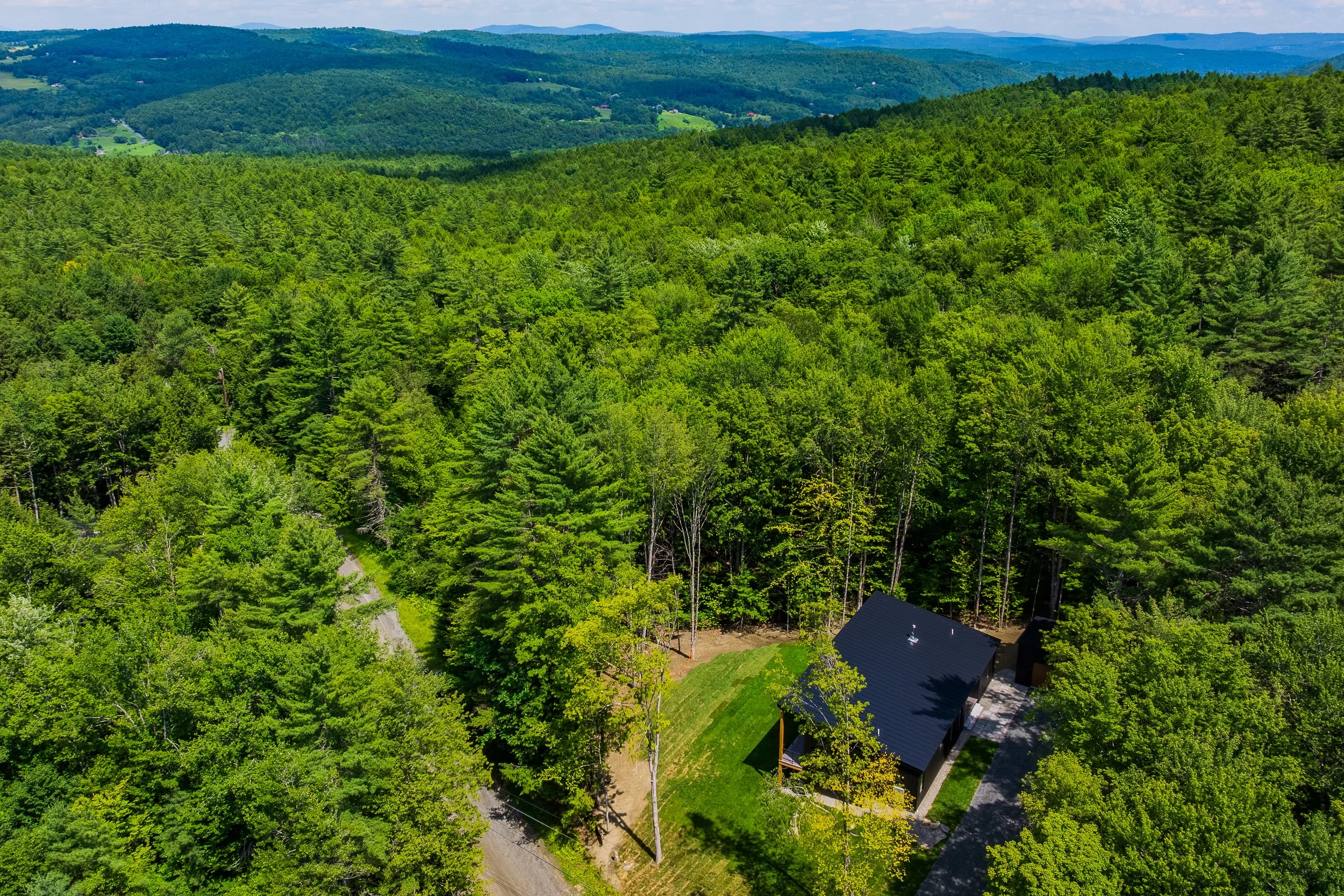 Aerial view of a modern cabin surrounded by dense green forest, with rolling mountains and rural valleys in the distance.