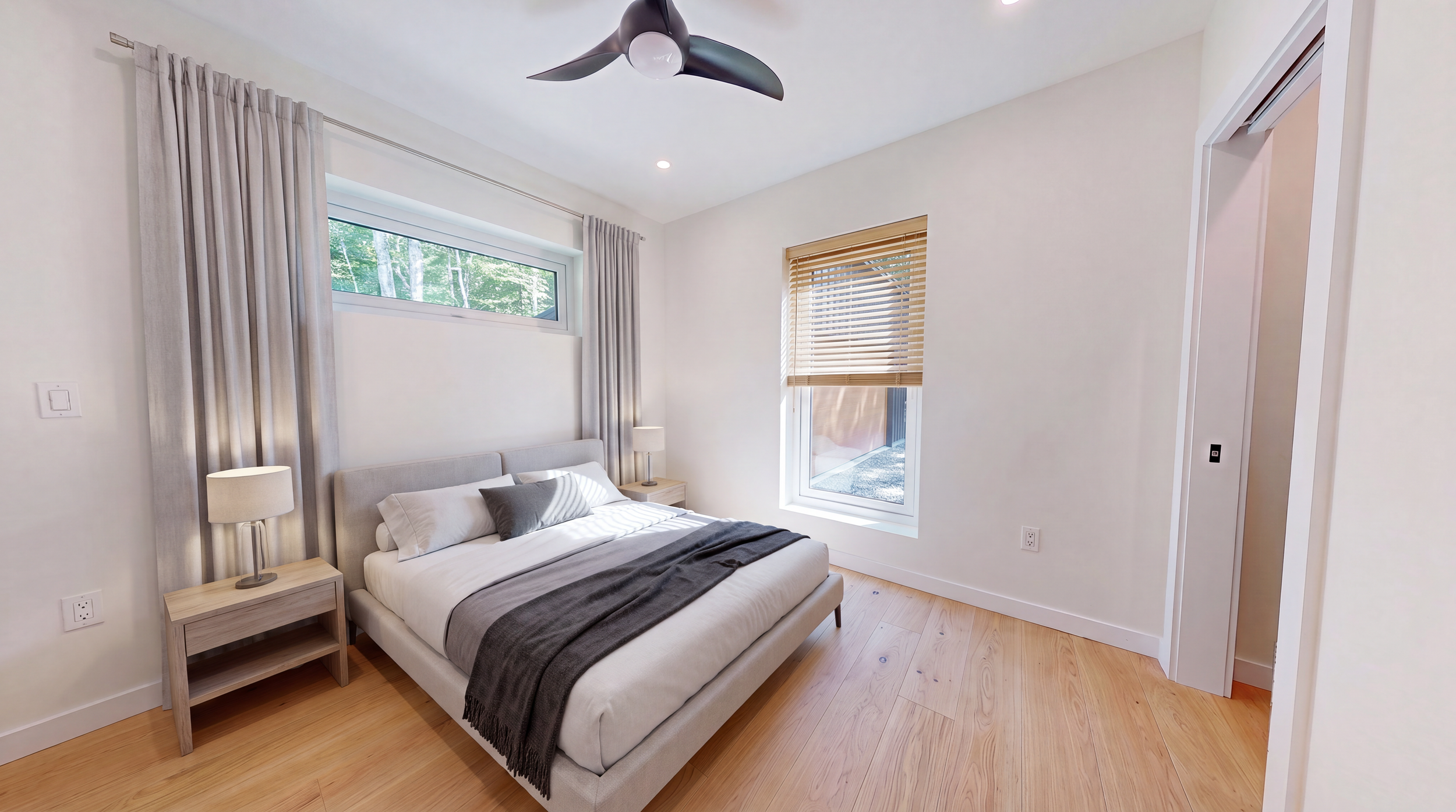 Modern bedroom with a neatly made bed, gray throw blanket, bedside tables and lamps, wood floors, ceiling fan, and large curtained windows.