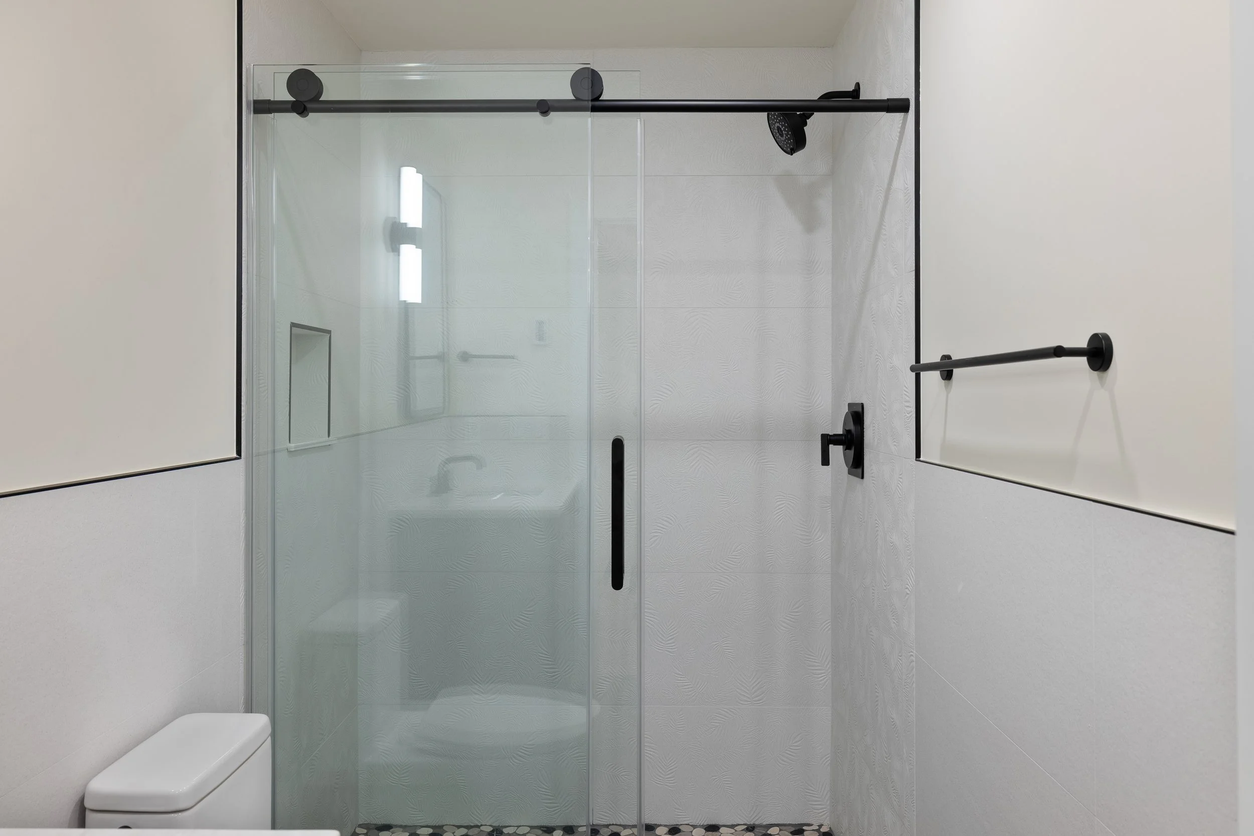 Modern bathroom with a clear glass walk-in shower, black fixtures, textured white tile walls, and pebble shower floor