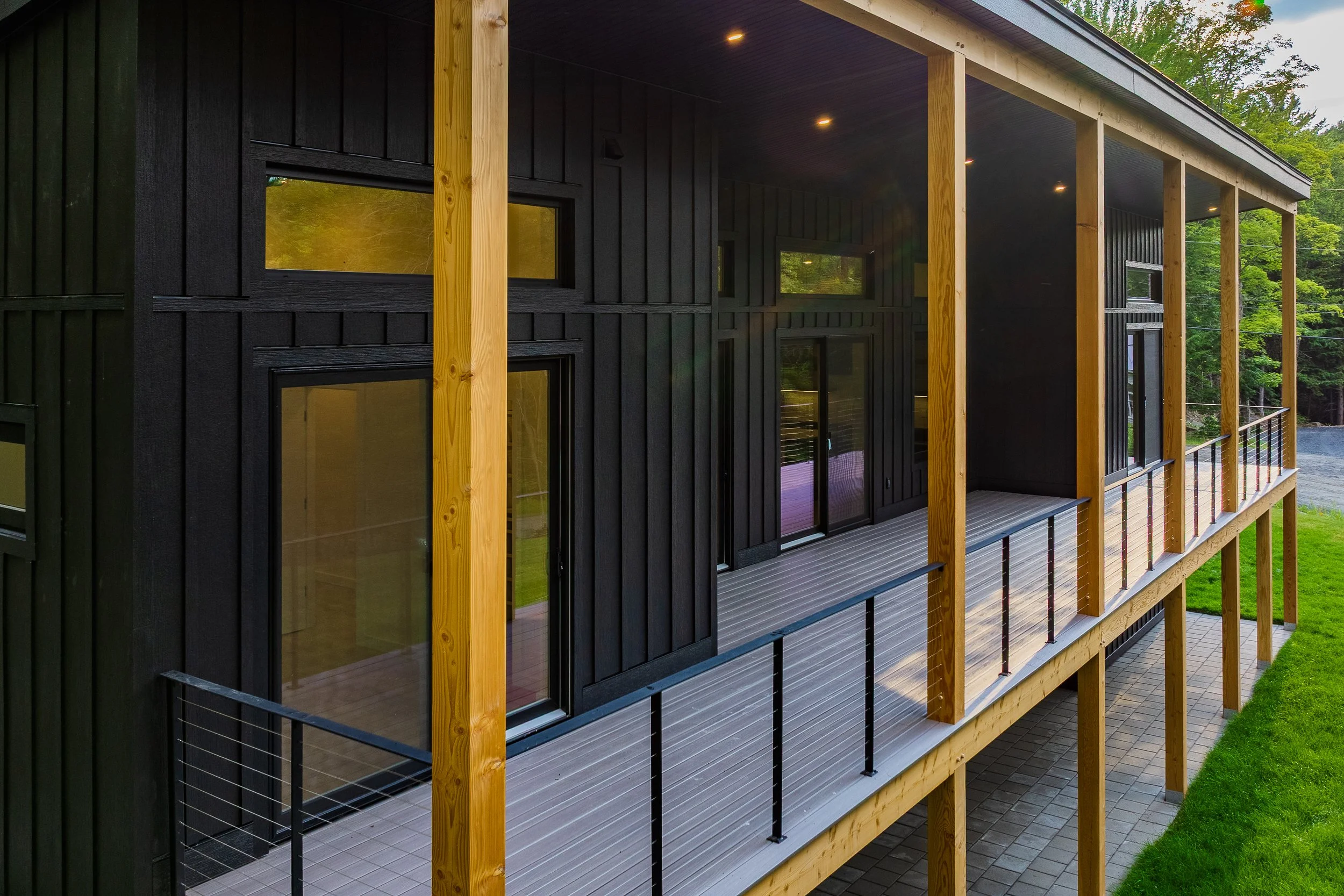 Modern black house with a covered wood deck, natural timber posts, cable railing, and forest surroundings