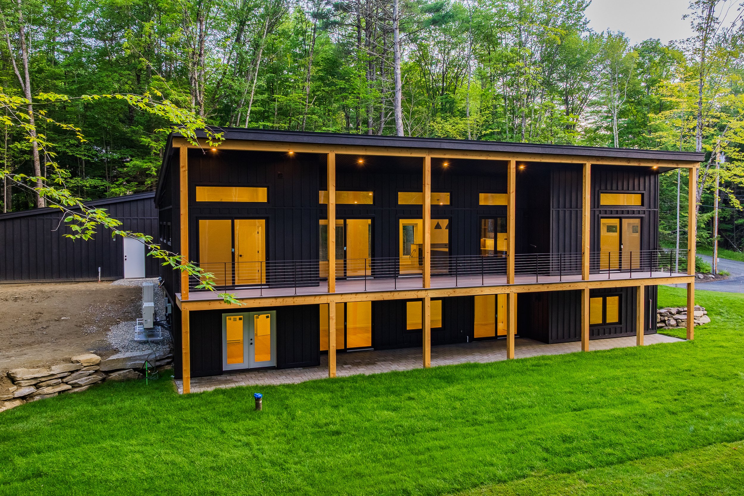 Modern black two-story home with warm wood accents, covered deck, and glowing windows in a forested setting