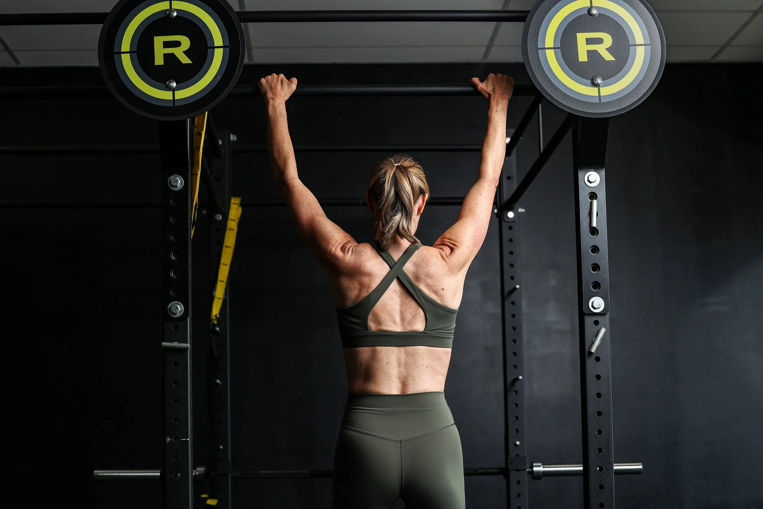 Stacie Carney wearing a green Vuori workout outfit while performing a pull up on a gym rack.