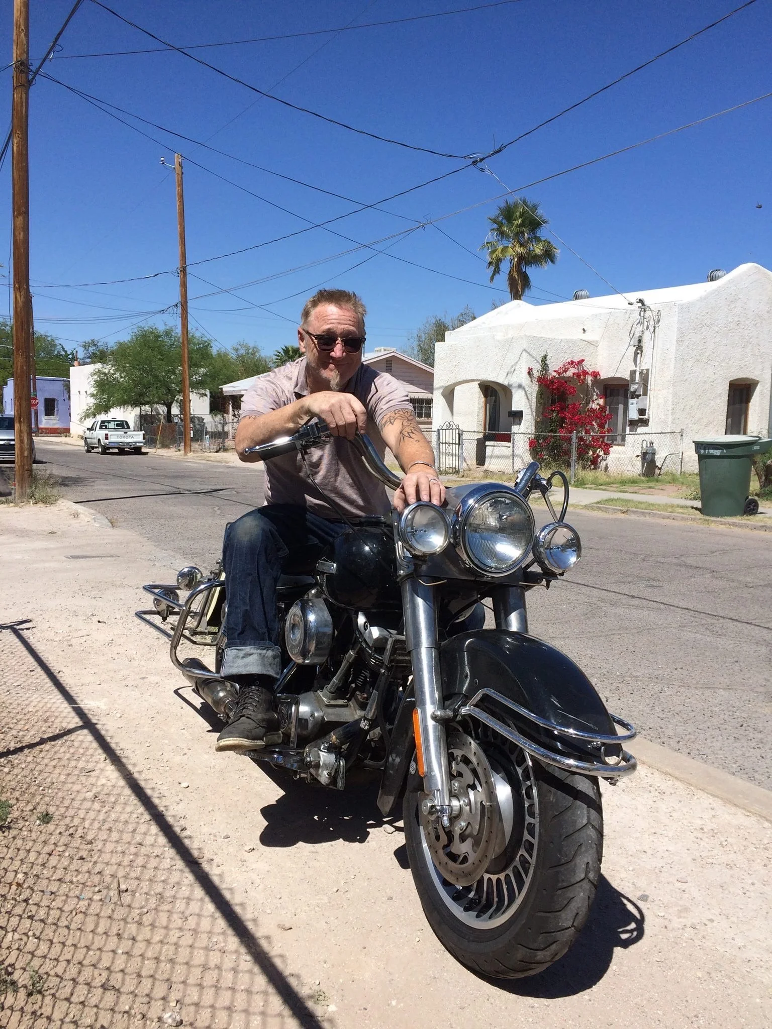 Fine artist Danny Perkins wearing sunglasses and cuffed jeans, seated on a black cruiser motorcycle outside his studio neighborhood in Tucson, Arizona