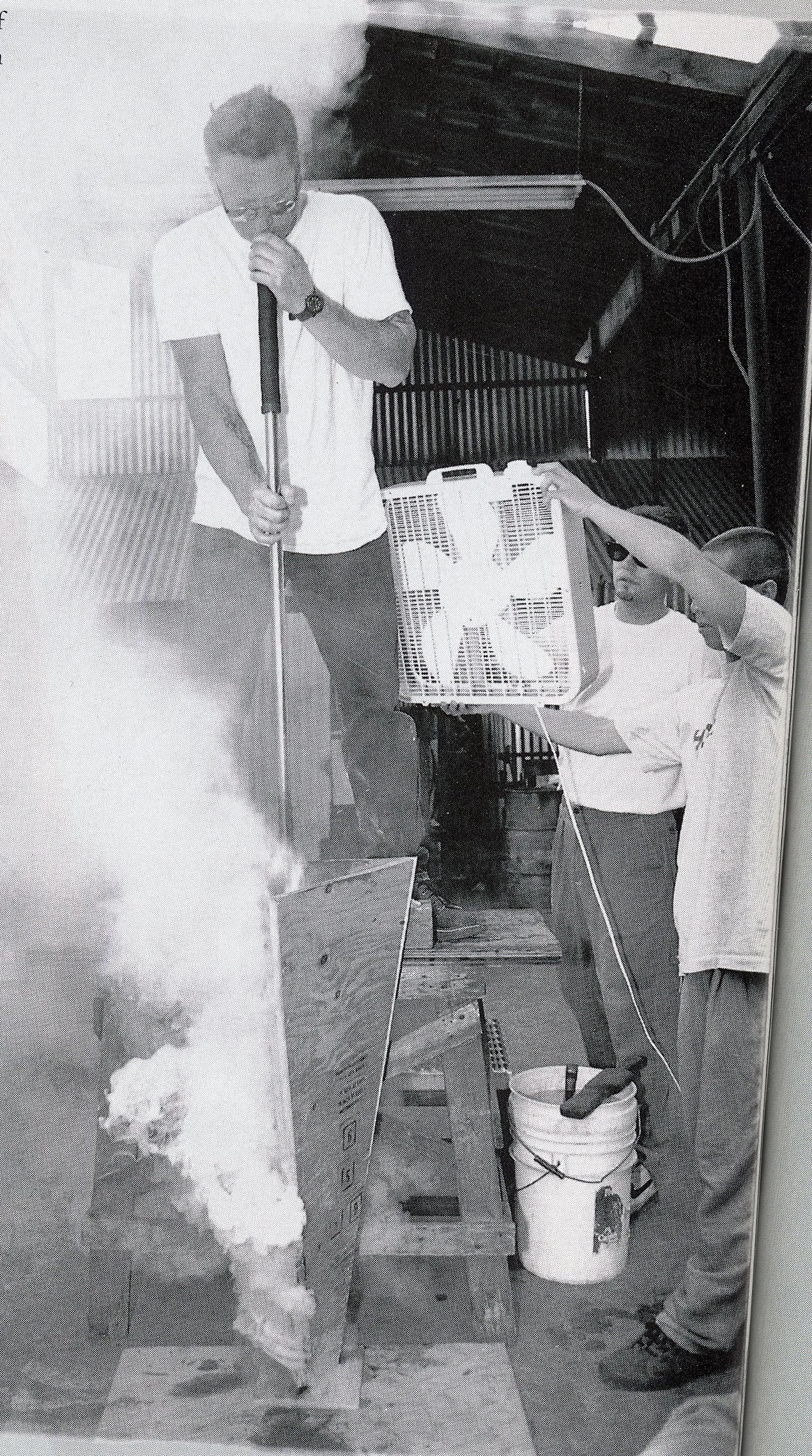 Black and white historical photograph of artist Danny Perkins standing atop a wooden platform, blowing into a long metal glassblowing pipe. Below, the glowing molten glass is lowered into a tall triangular wooden mold emitting heat or steam