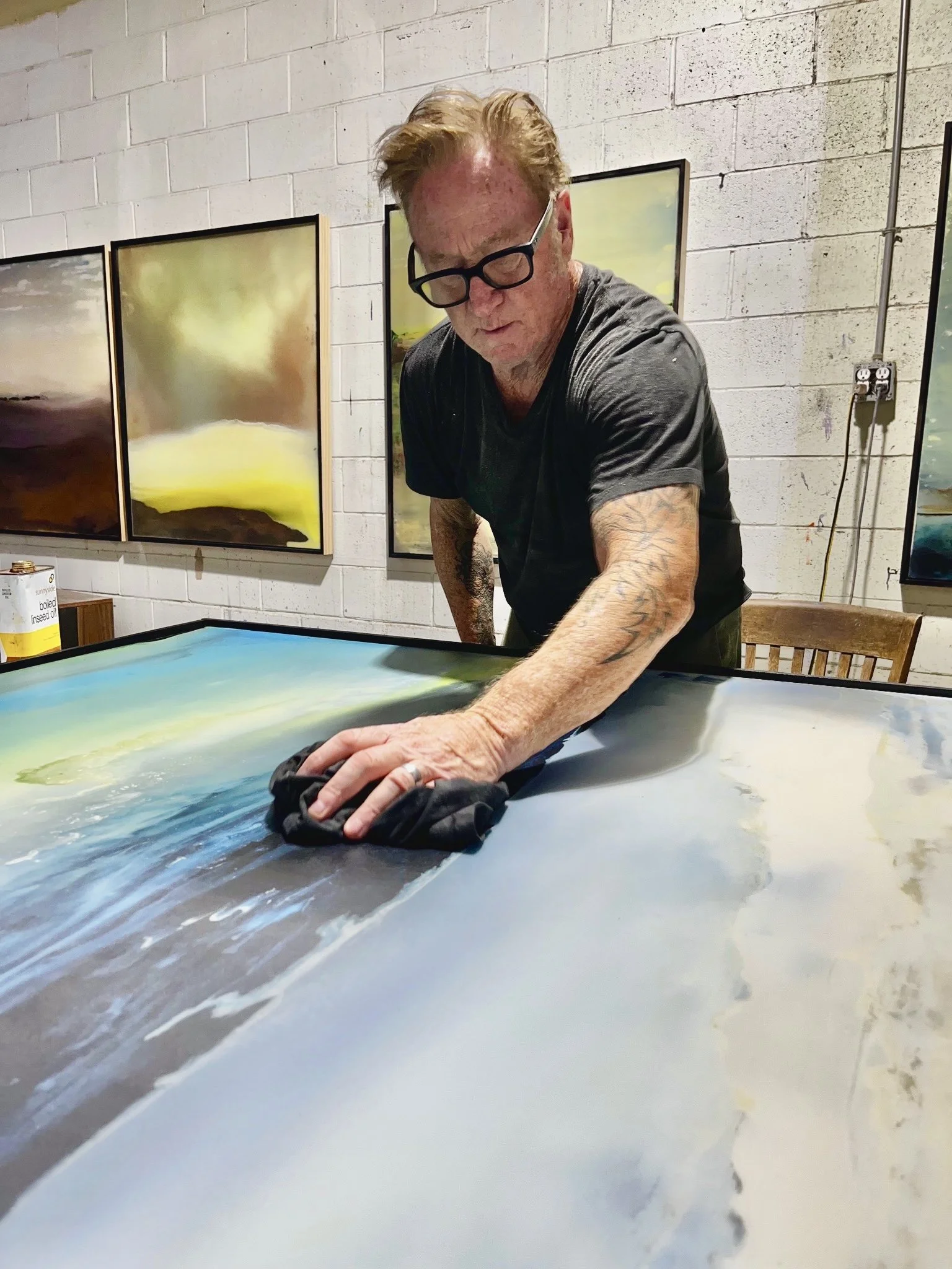 Artist Danny Perkins hand-finishing a large oil on birch panel work with resin in his cinder-block studio, with completed 2025 color field paintings mounted on the wall behind him.