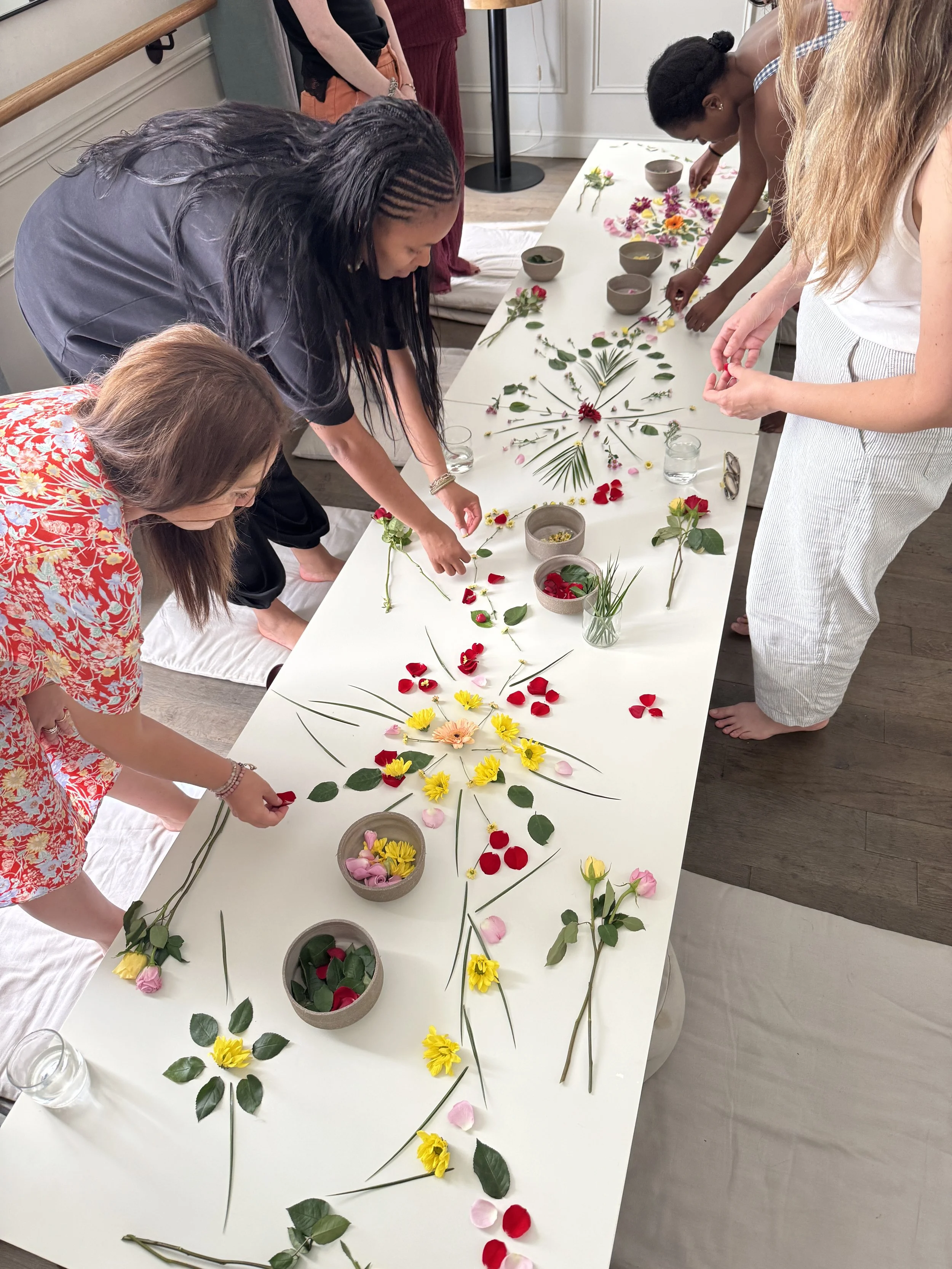 People create floral arrangements with fresh flowers, leaves, petals, and stems on a long white table.