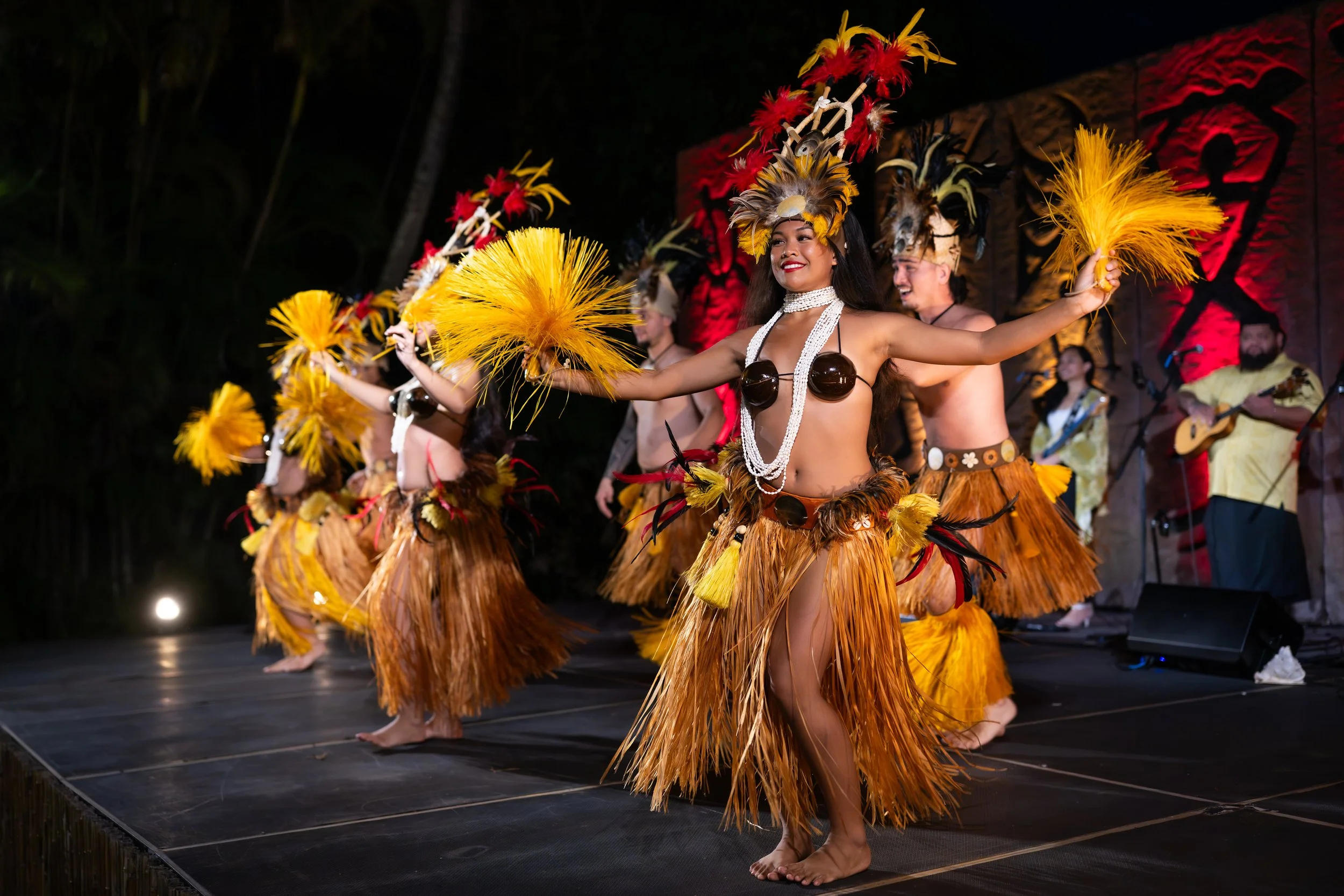 Tahitian dancers performing onstage in grass skirts and colorful feathered headdresses.