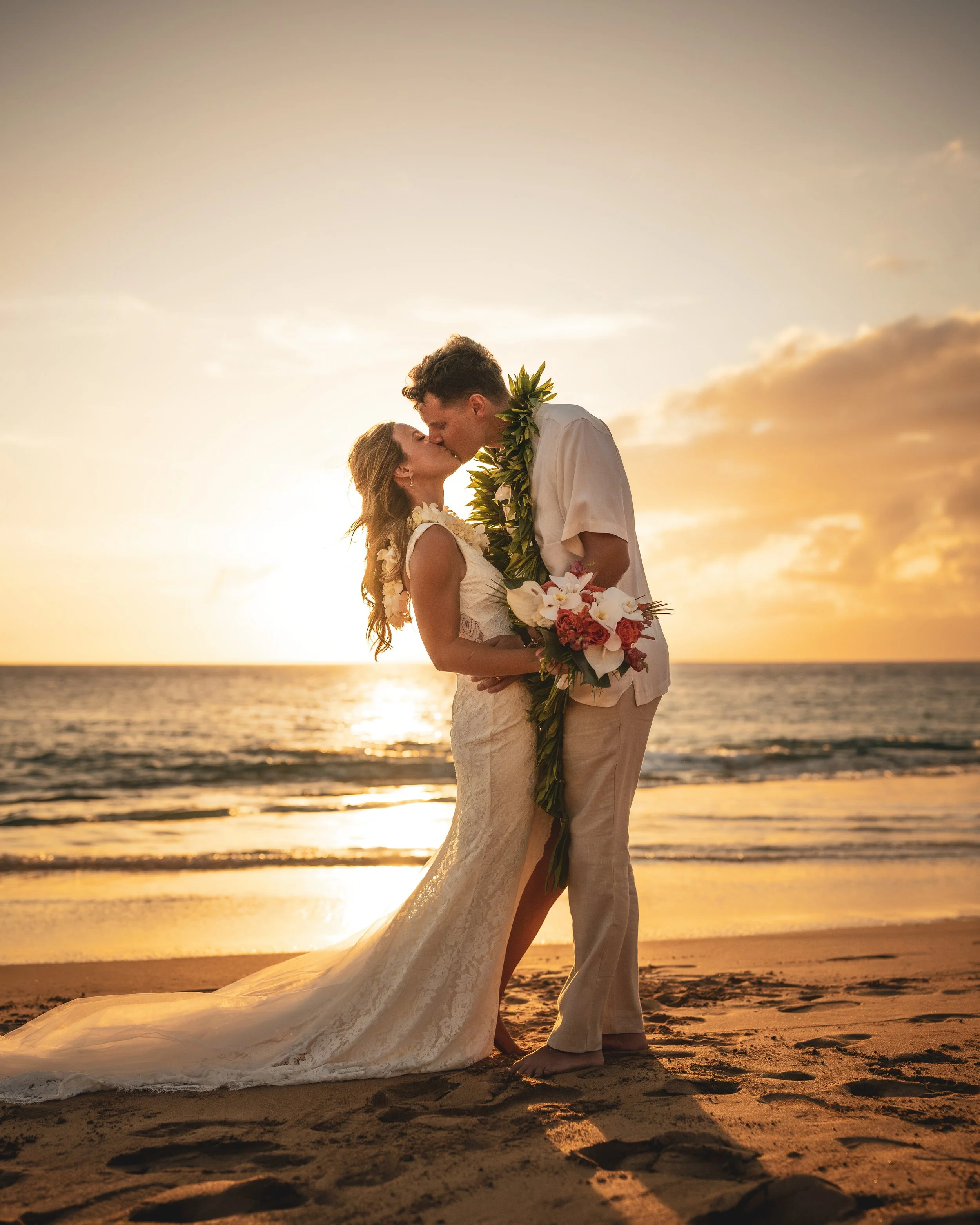 Newlywed couple kissing on a beach at sunset, wearing leis and holding a tropical wedding bouquet.