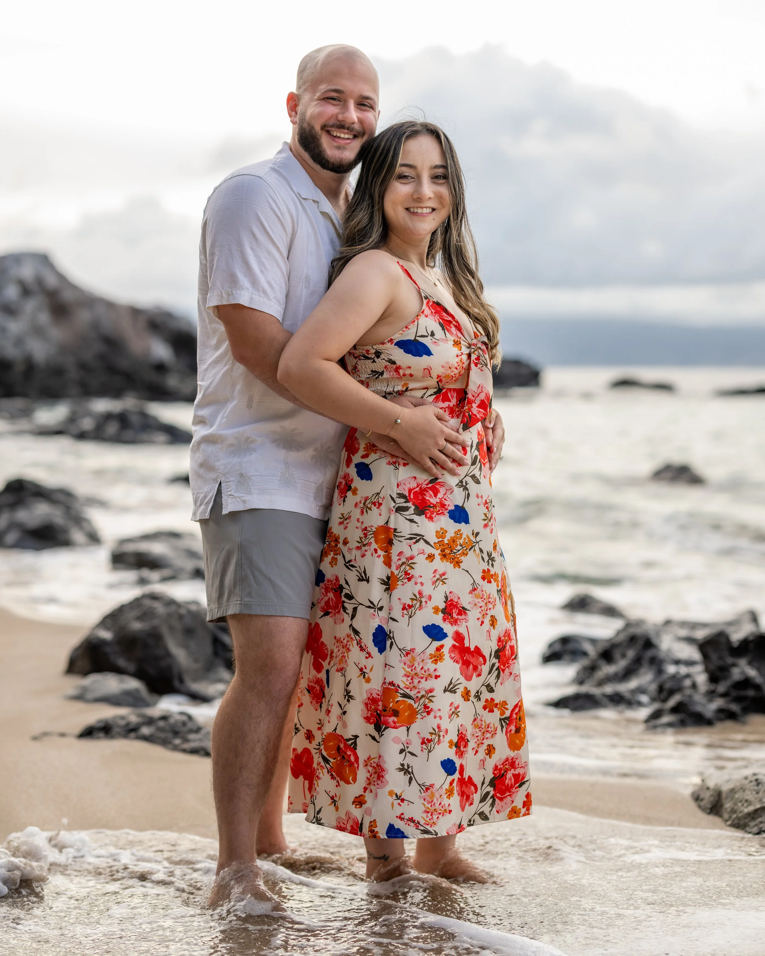 Couple embracing on a rocky Maui beach, the woman wearing a colorful floral dress.