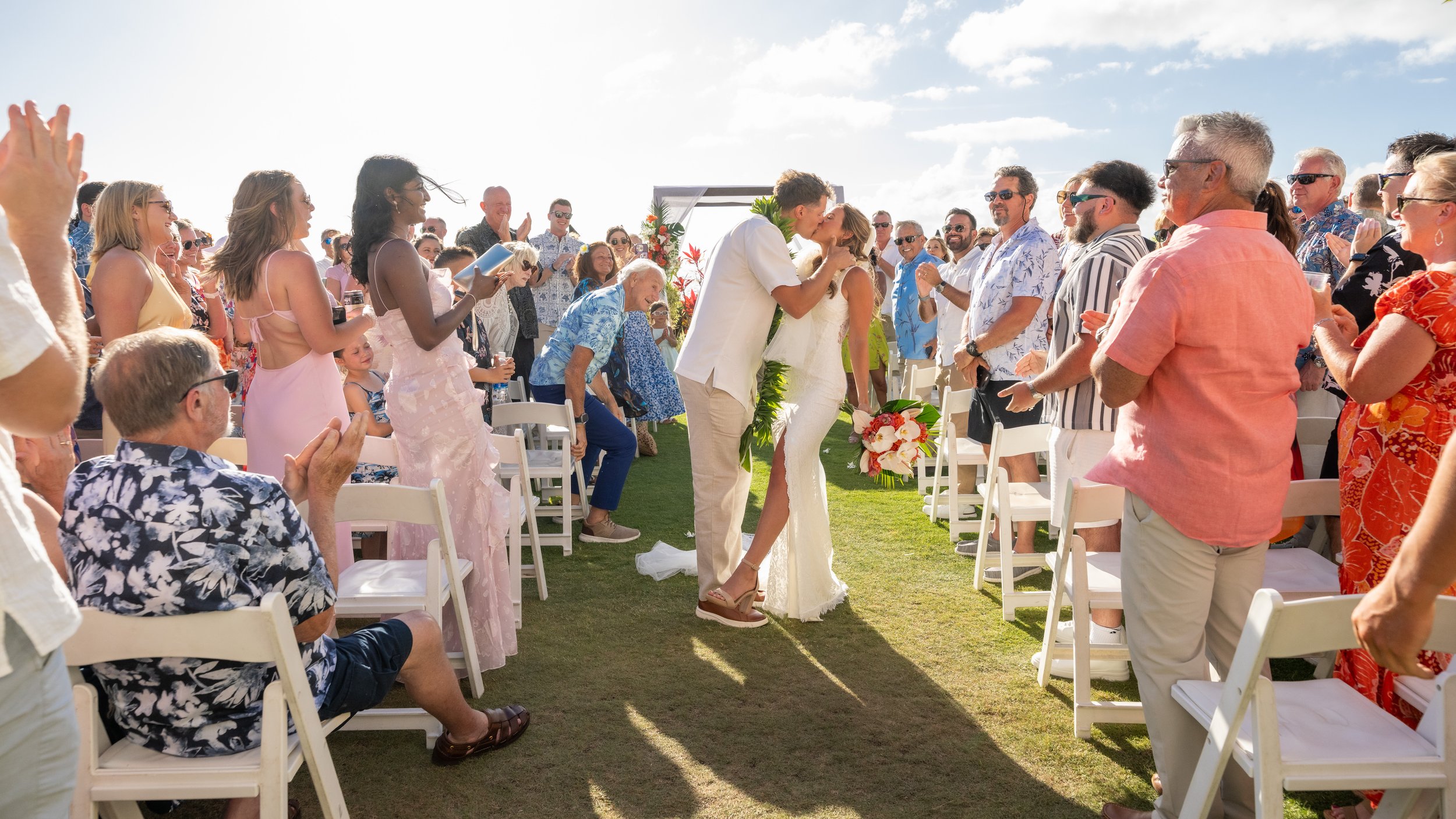 Bride and groom kissing in the aisle as wedding guests applaud at an outdoor Maui ceremony.