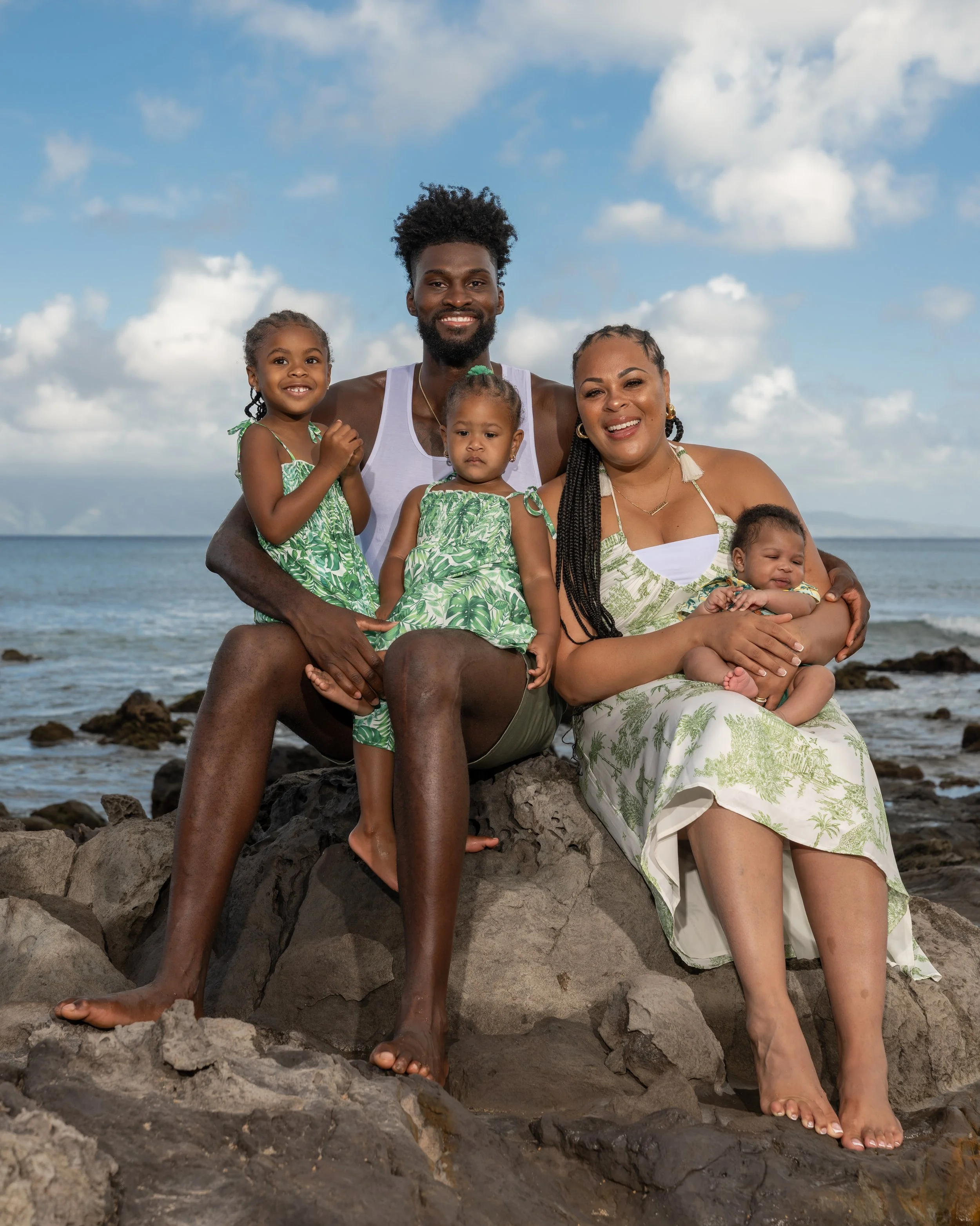 Mom and dad with their three children, gathered on coastal rocks with the ocean behind them.