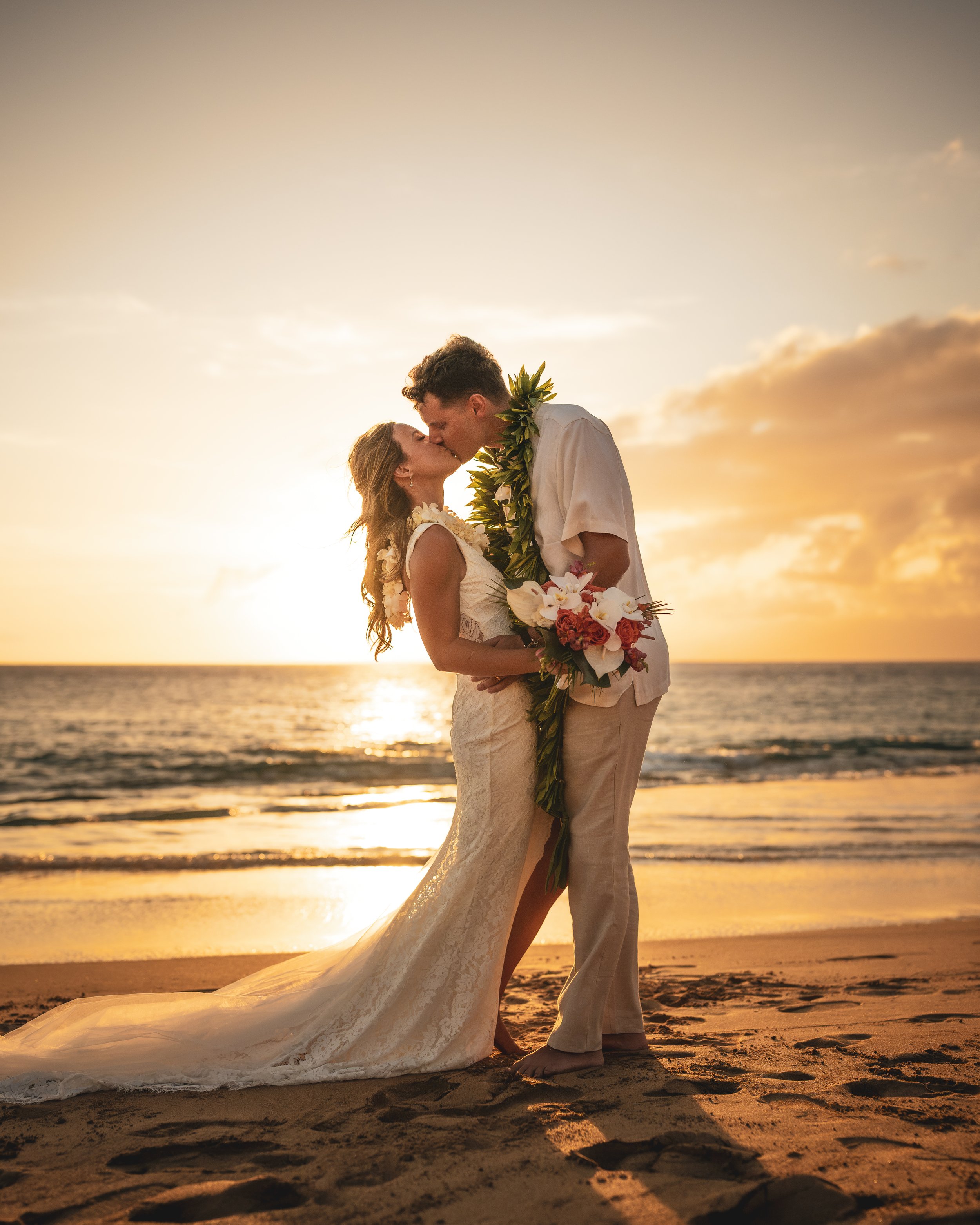 Bride and groom kissing on a Maui beach at sunset, with her wedding dress train trailing across the sand and golden light over the ocean.