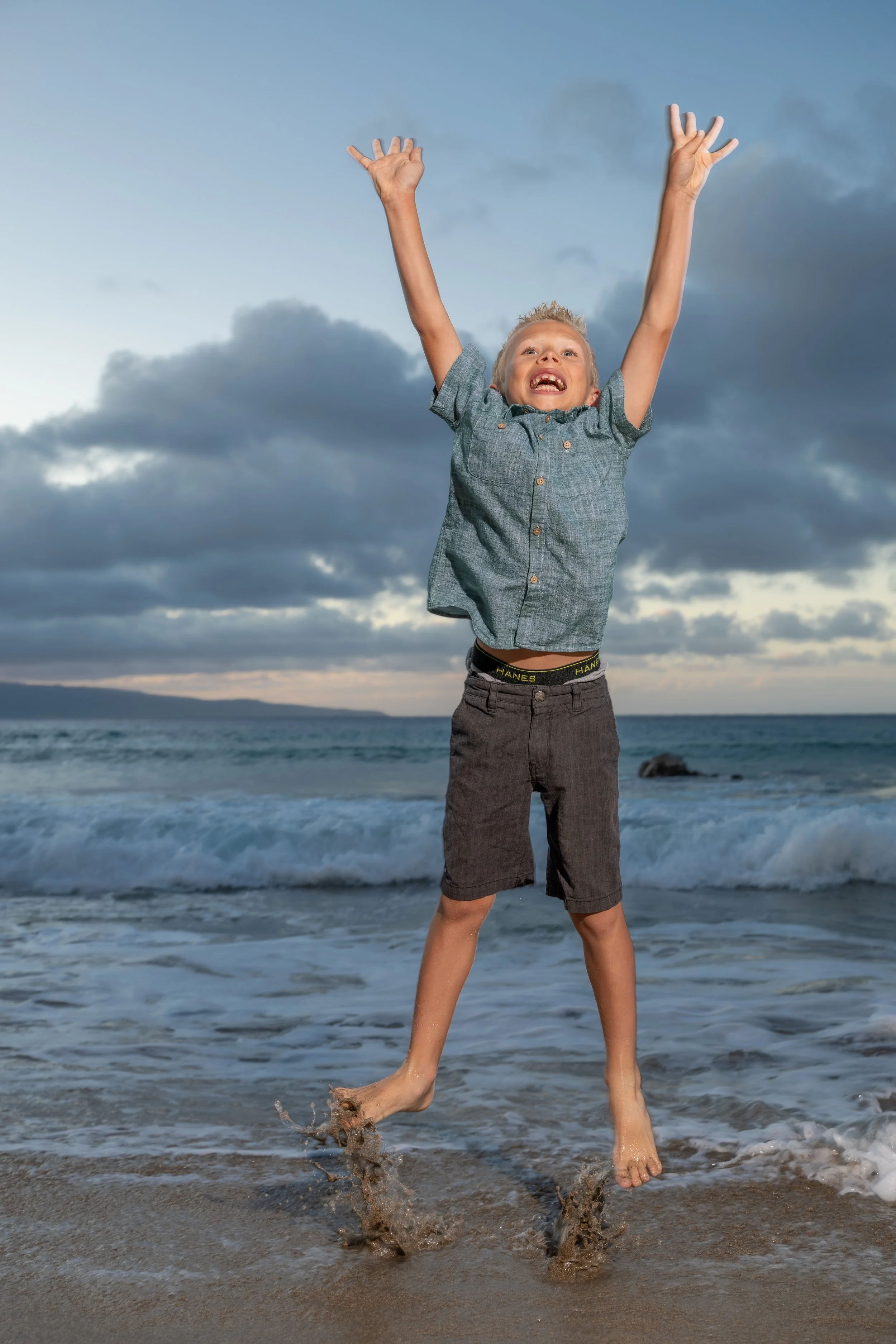 Young boy jumping with his arms raised as waves splash around his feet on a Maui beach.