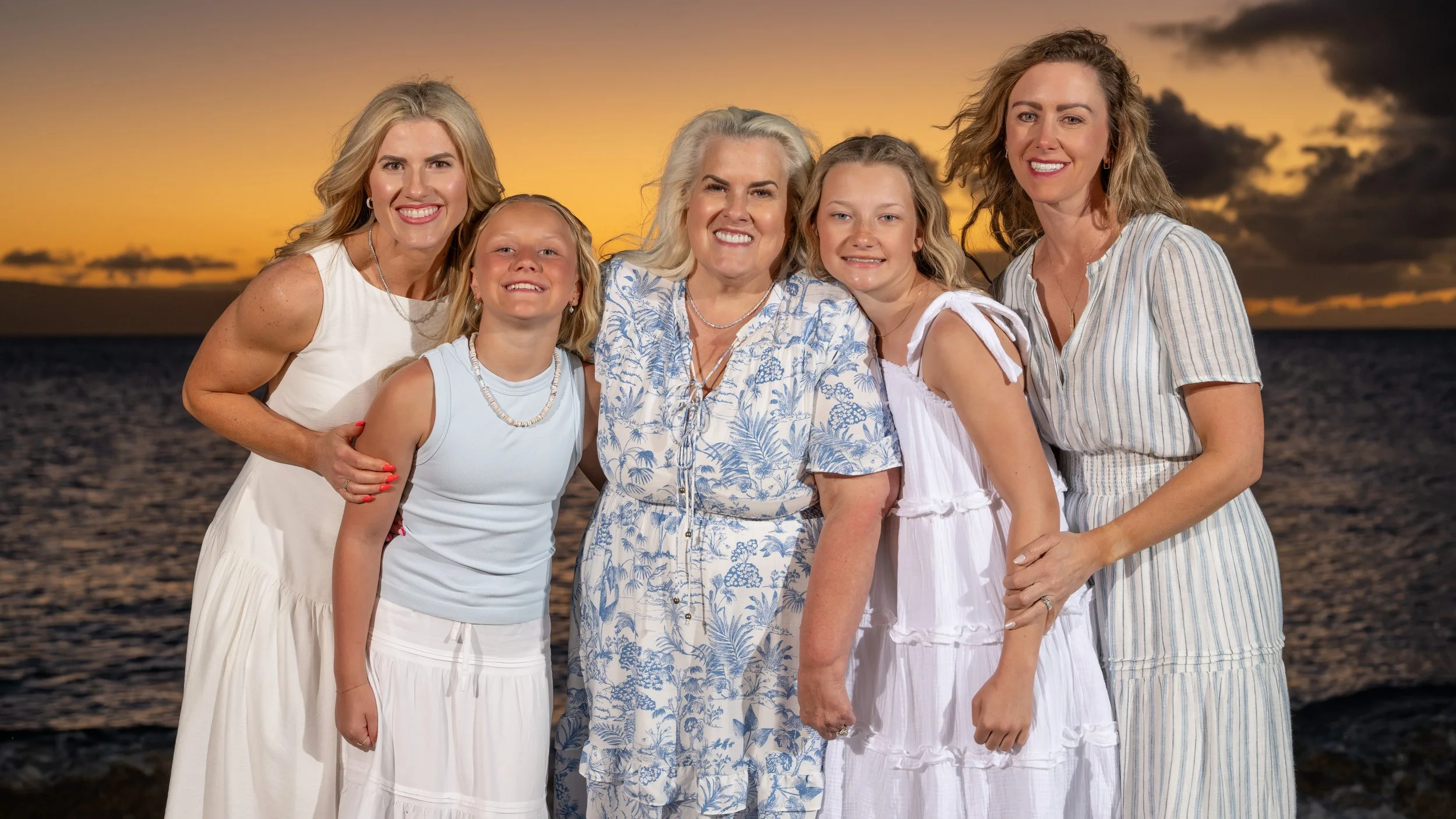 Five family members gathered closely together on a Maui beach, smiling against a golden sunset over the ocean.