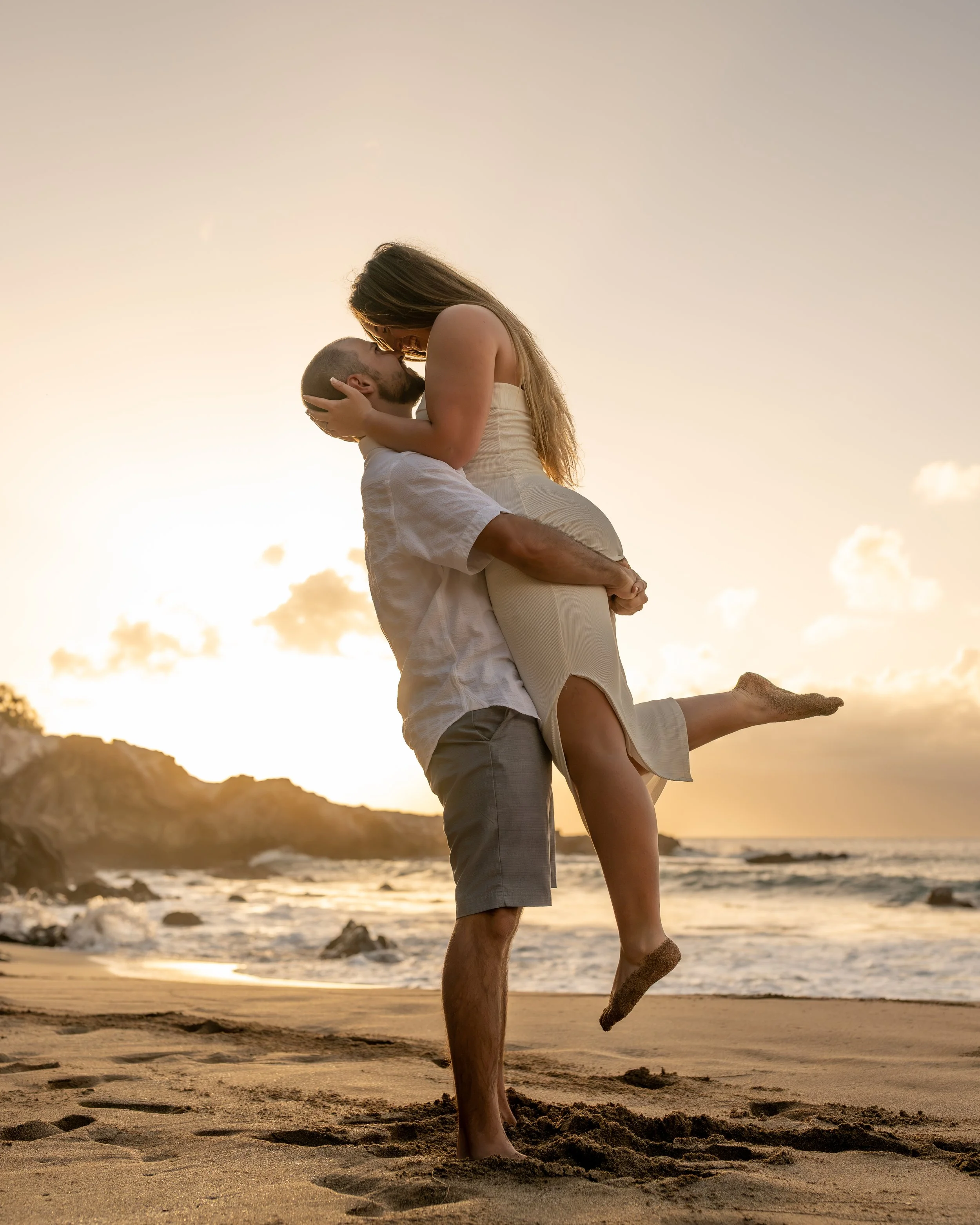 Couple embracing on the beach at sunset, one lifting the other off the sand.