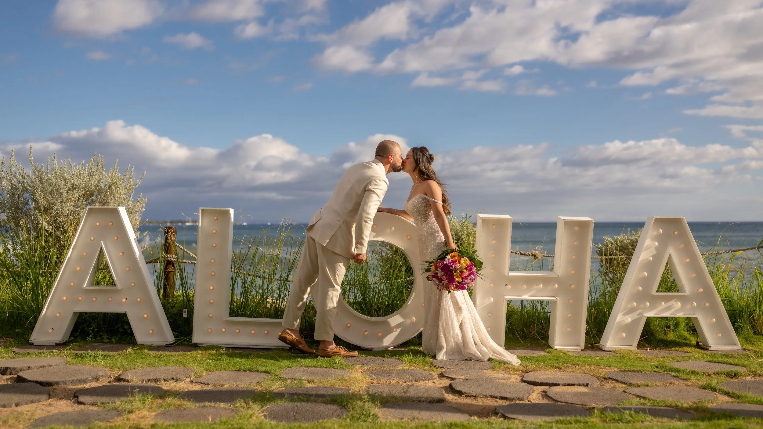 Bride and groom leaning in for a kiss beside a white fence overlooking the ocean, with blue skies above Maui.