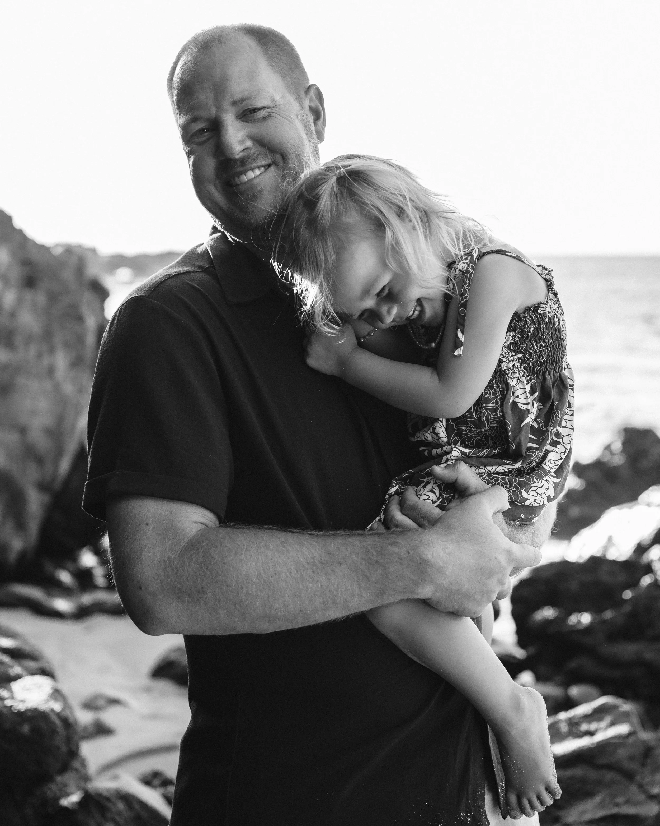 Black-and-white portrait of a father holding his laughing daughter on a Maui beach.