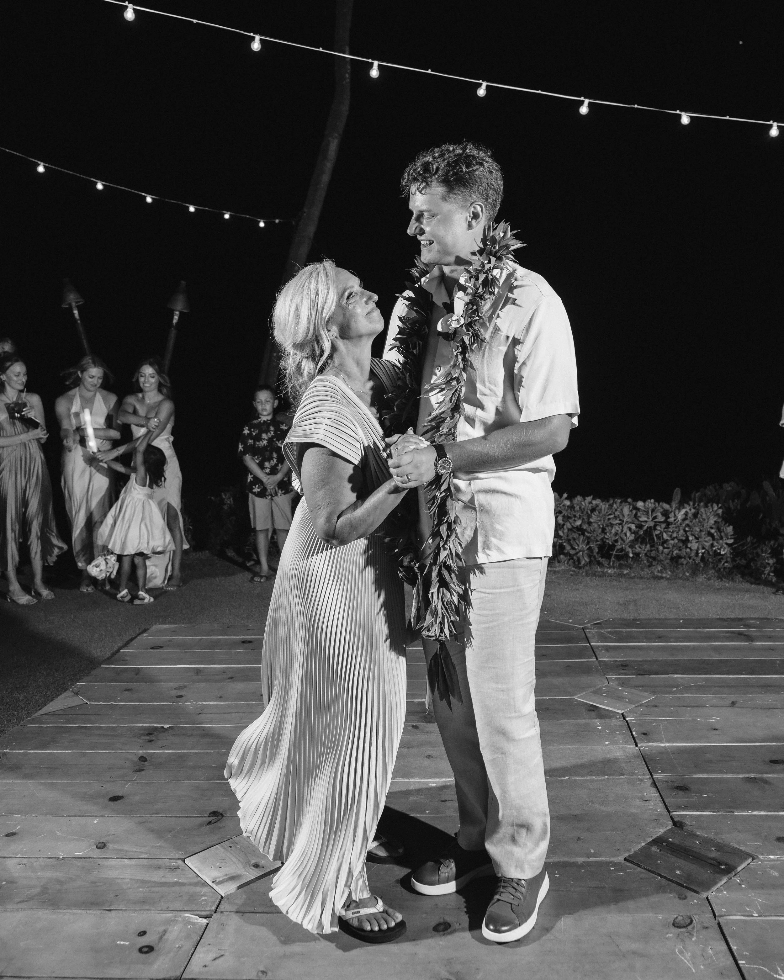Black-and-white photograph of the groom dancing with a woman beneath string lights at an outdoor Maui wedding reception.