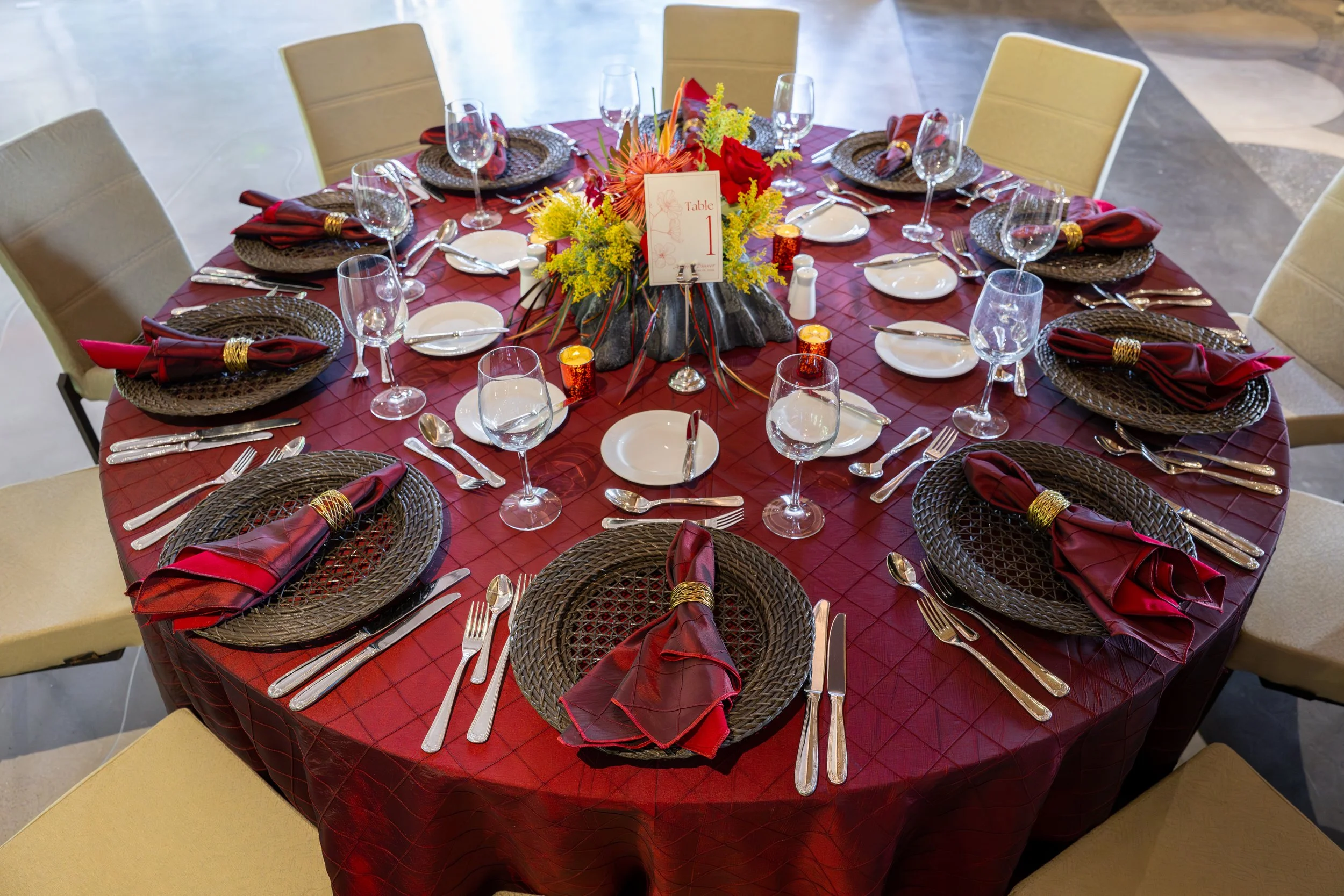 Table set for a corporate dinner with red linens, woven chargers, and a tropical floral centerpiece.