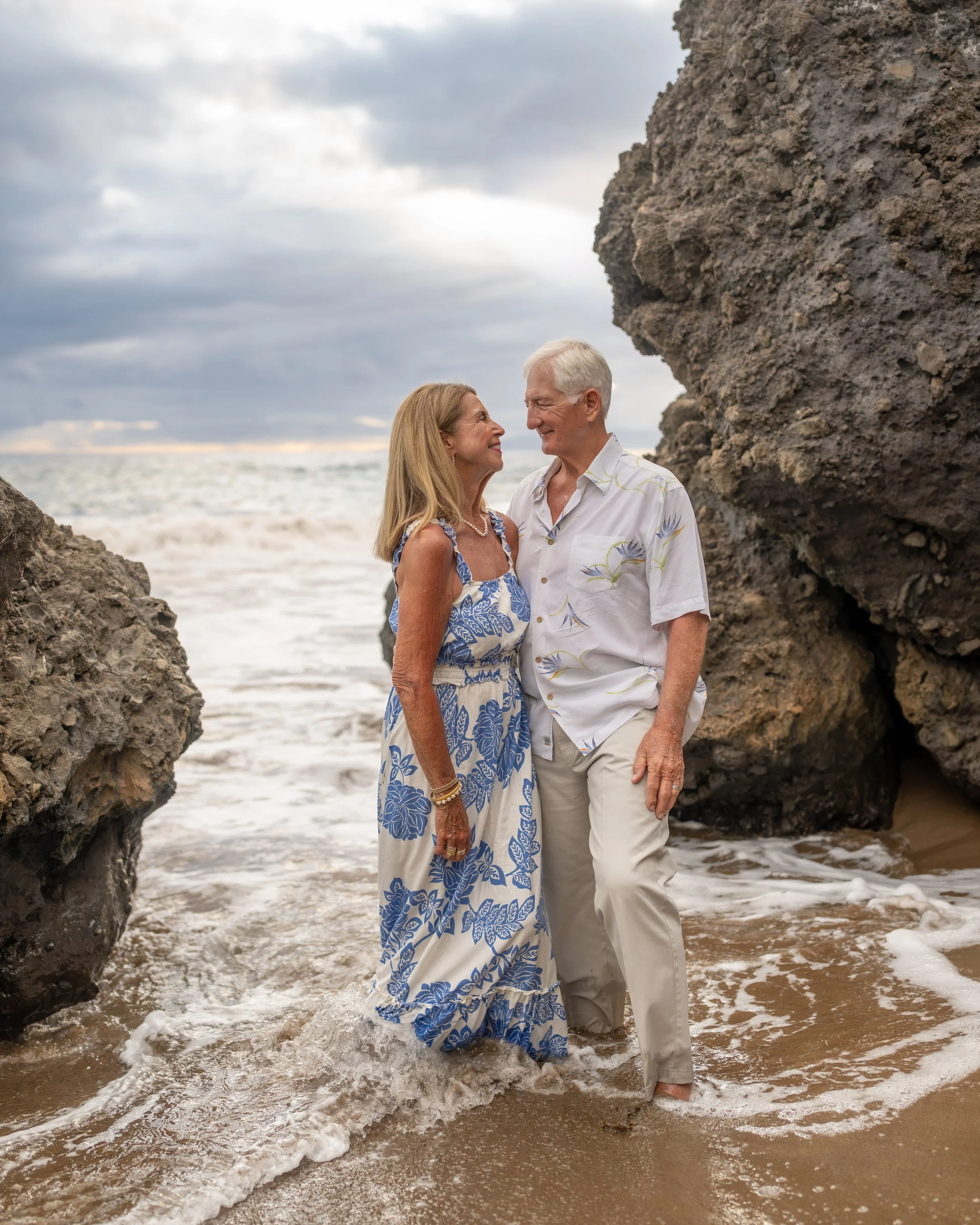 Couple holding hands at the water’s edge between large coastal rocks.