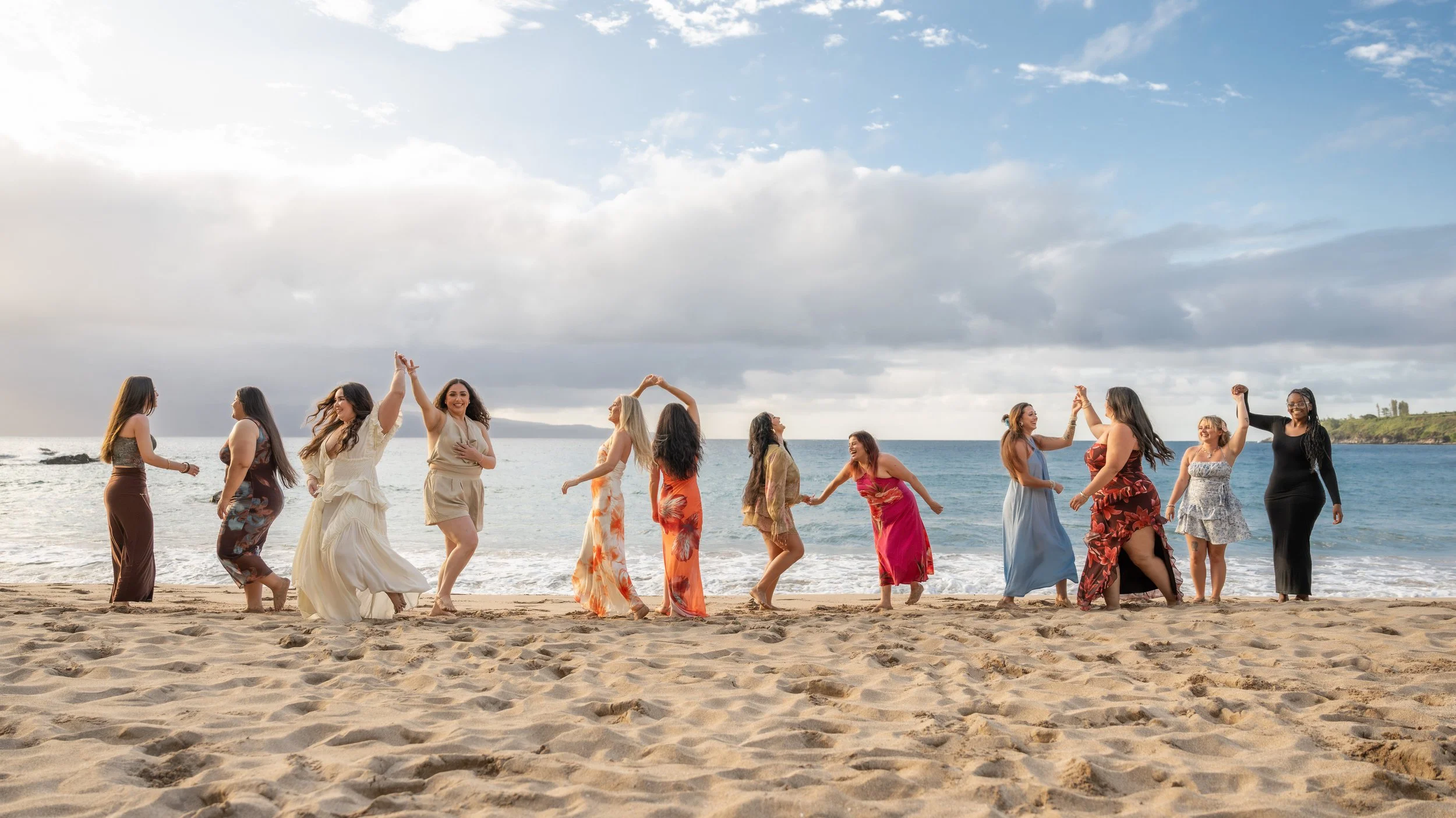 Family and friends holding hands and twirling along a Maui shoreline in colorful dresses.