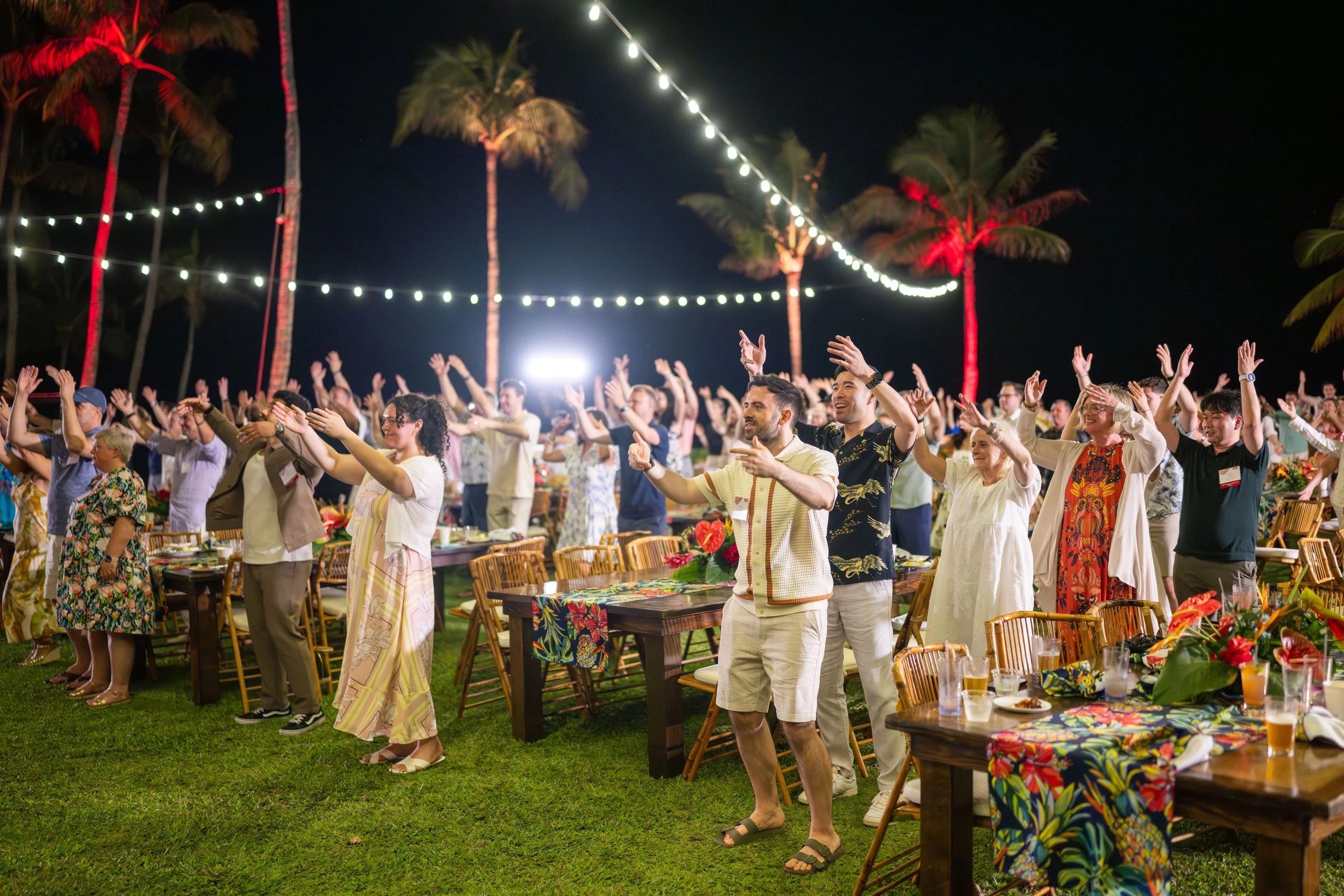 Guests learning hula with their arms raised beneath palm trees and string lights at an evening luau.