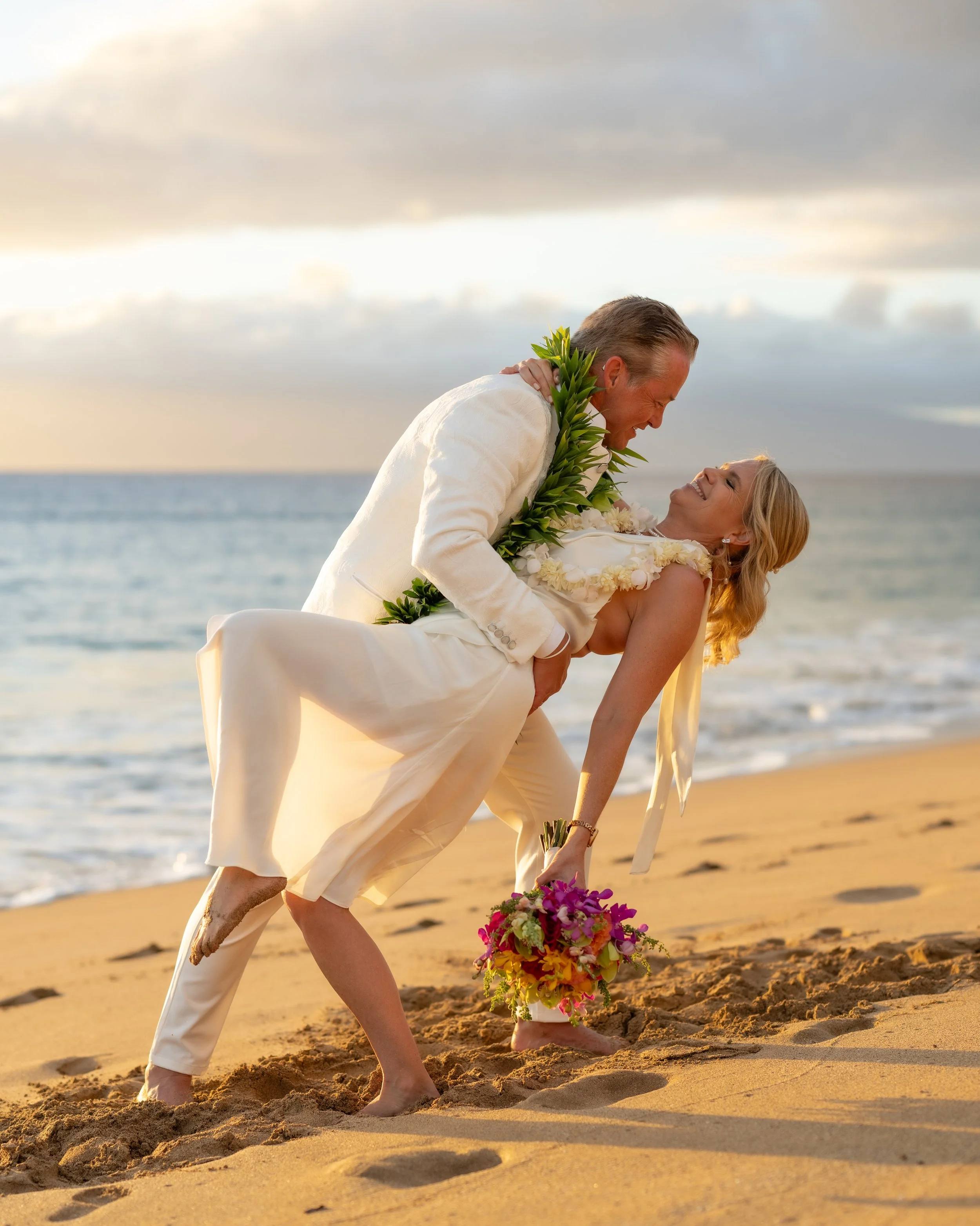 Groom dipping the bride for a kiss on a Maui beach, her colorful bouquet held near the sand.
