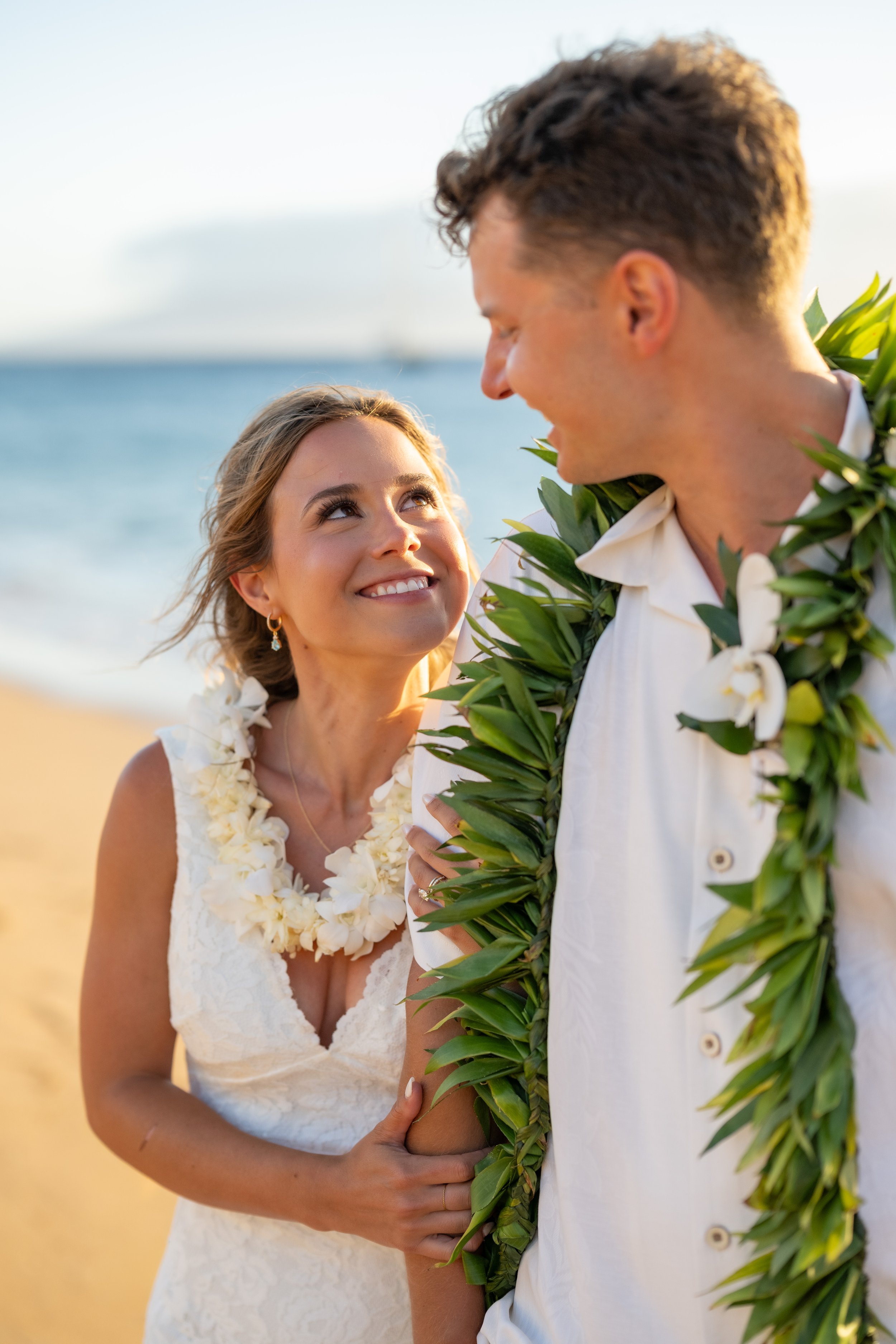 Bride smiling up at the groom as he looks down at her, both wearing leis on a Maui beach.