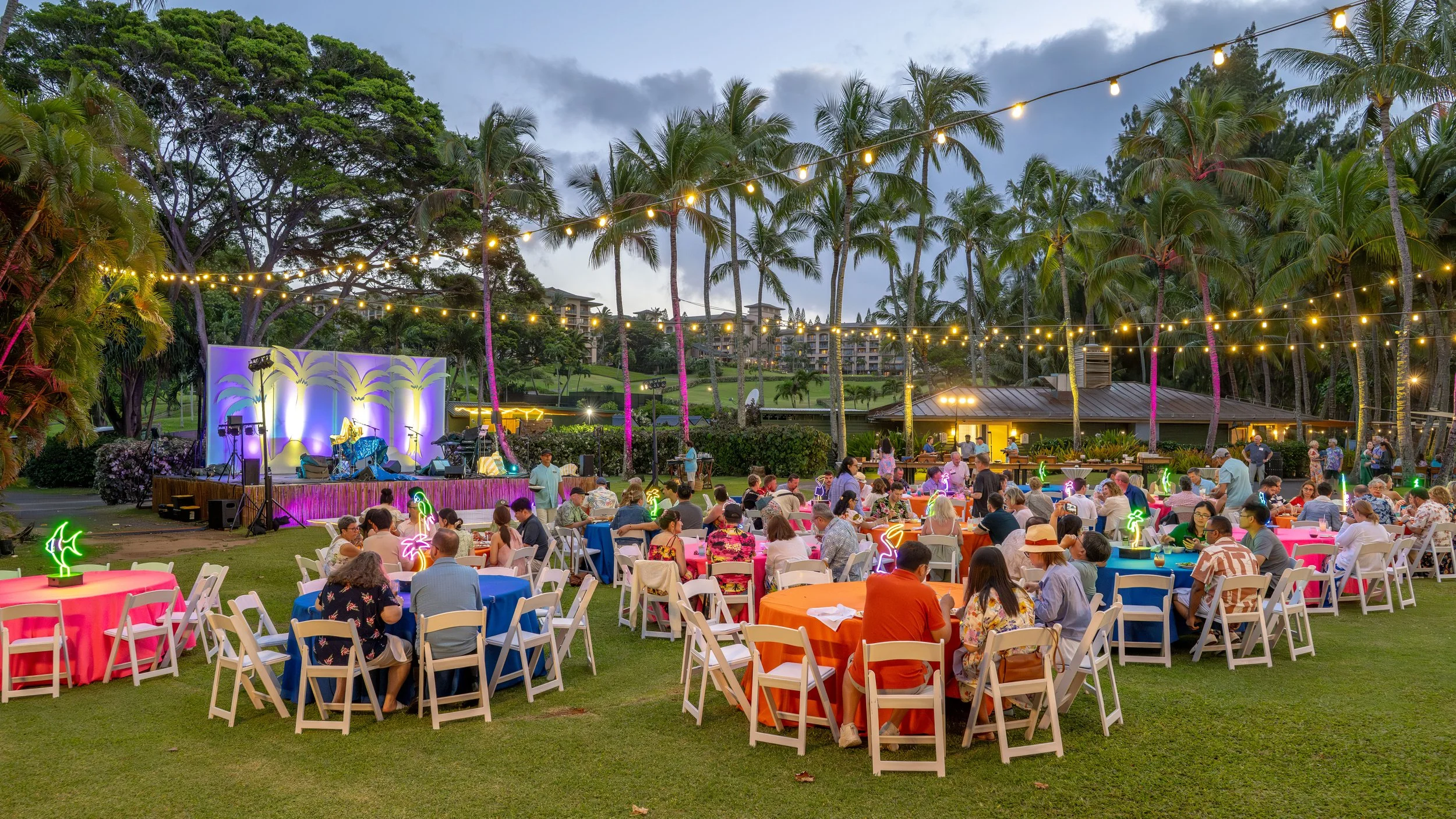 Outdoor corporate dinner with colorful seating, palm trees, string lights, and a stage.