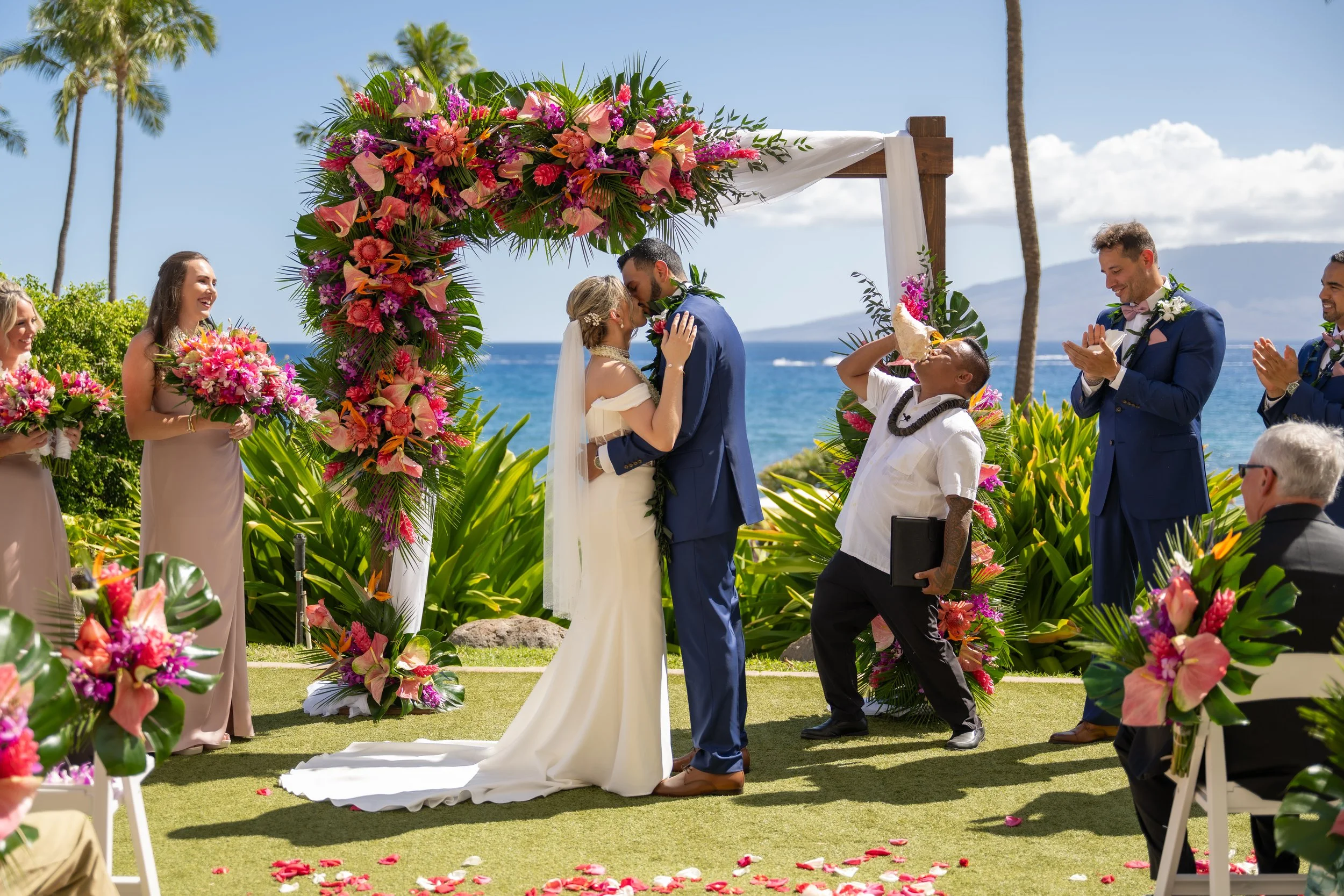 Bride and groom kissing beneath a colorful floral arch during an outdoor wedding ceremony.