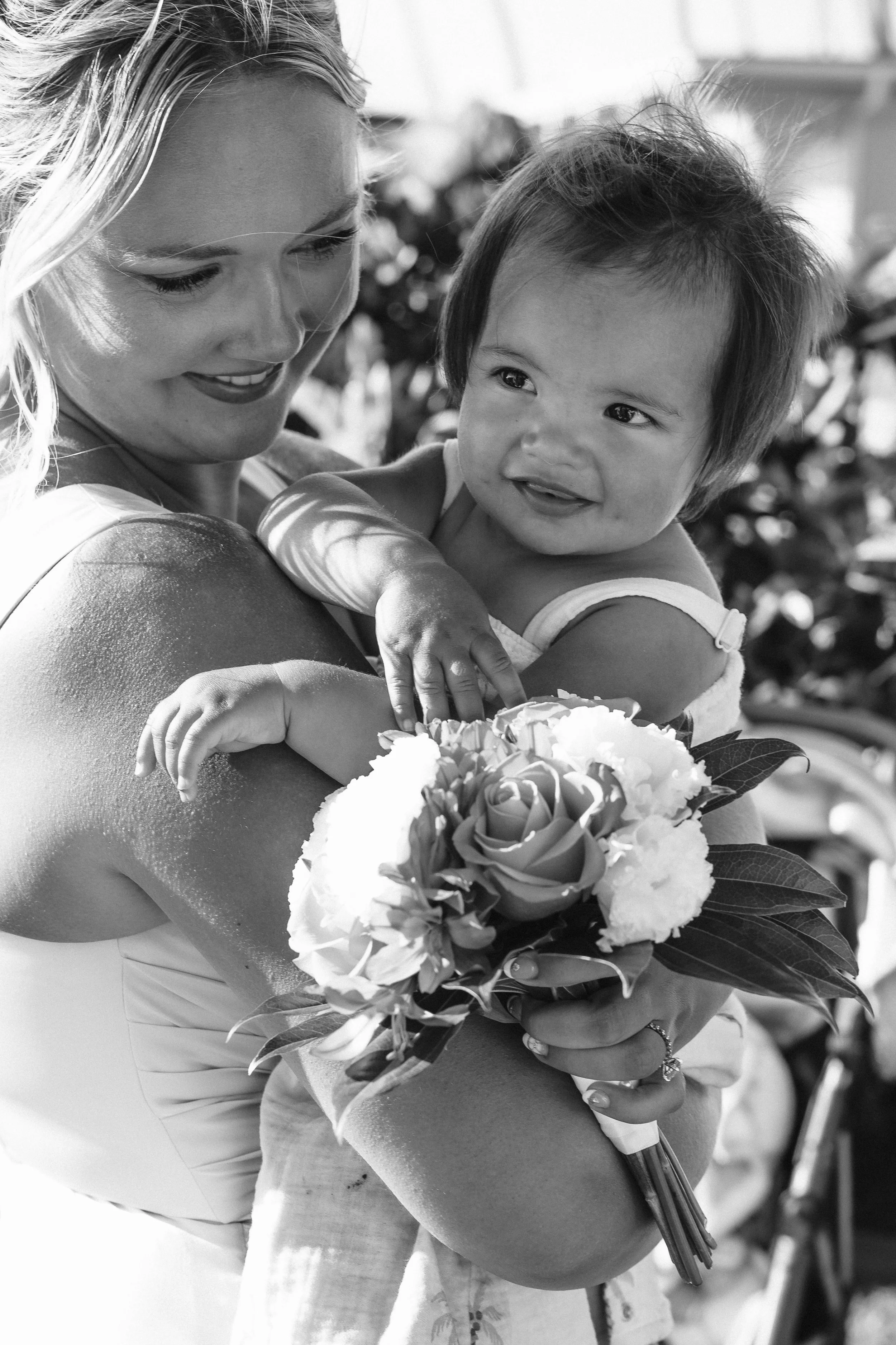 Black-and-white photograph of a bridesmaid holding a baby and a flower bouquet at an outdoor Maui wedding.