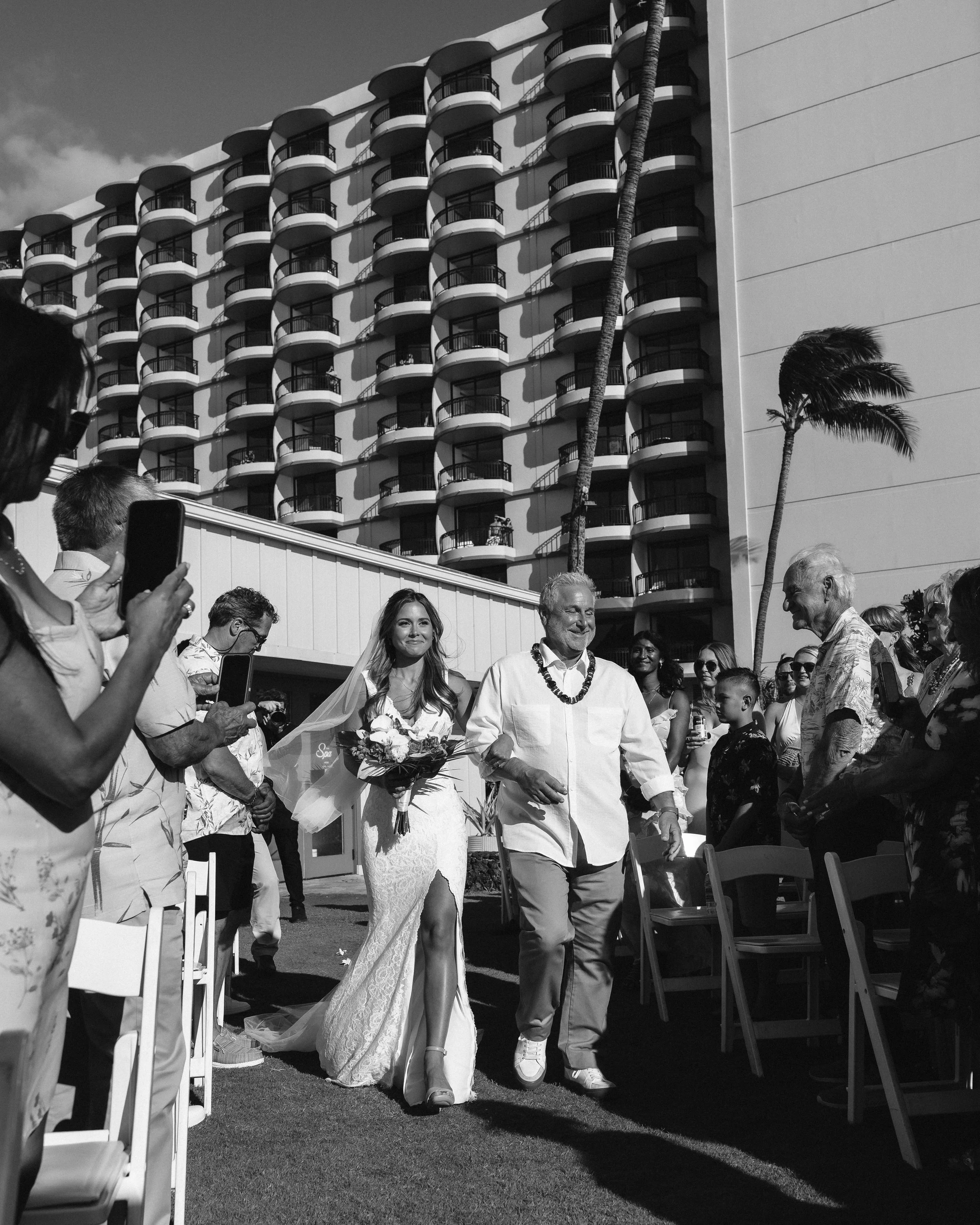 Black-and-white photograph of a bride walking down the aisle with her father as guests watch at an outdoor Maui wedding.