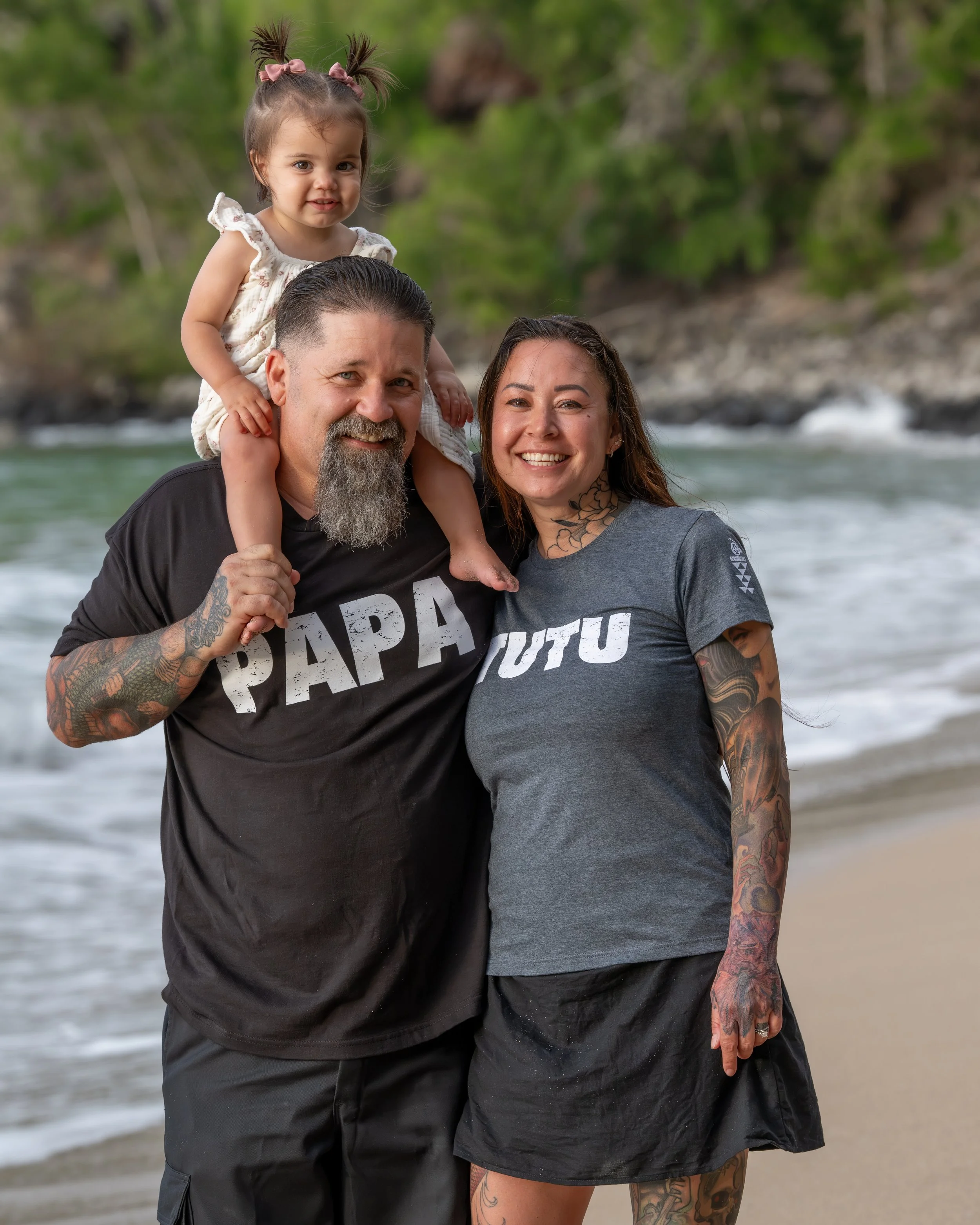 Smiling grandparents on a Maui beach with their grandchild riding on the grandfather’s shoulders, wearing shirts with Hawaiian words for grandfather and grandmother.