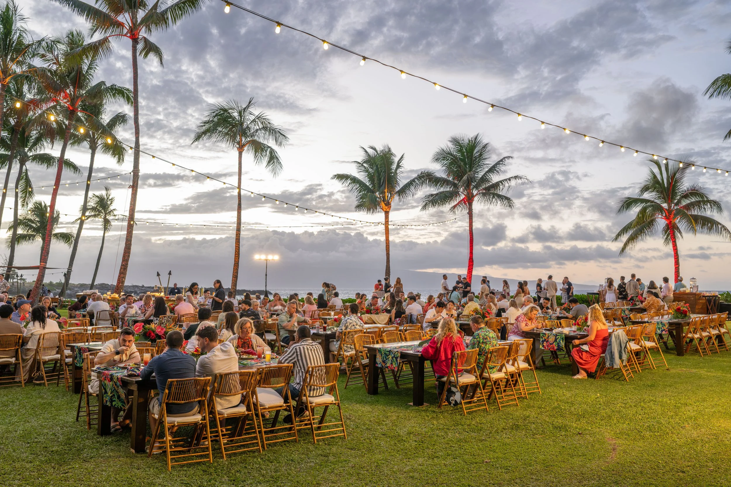 Guests dining on an oceanfront lawn beneath palm trees and string lights at dusk.