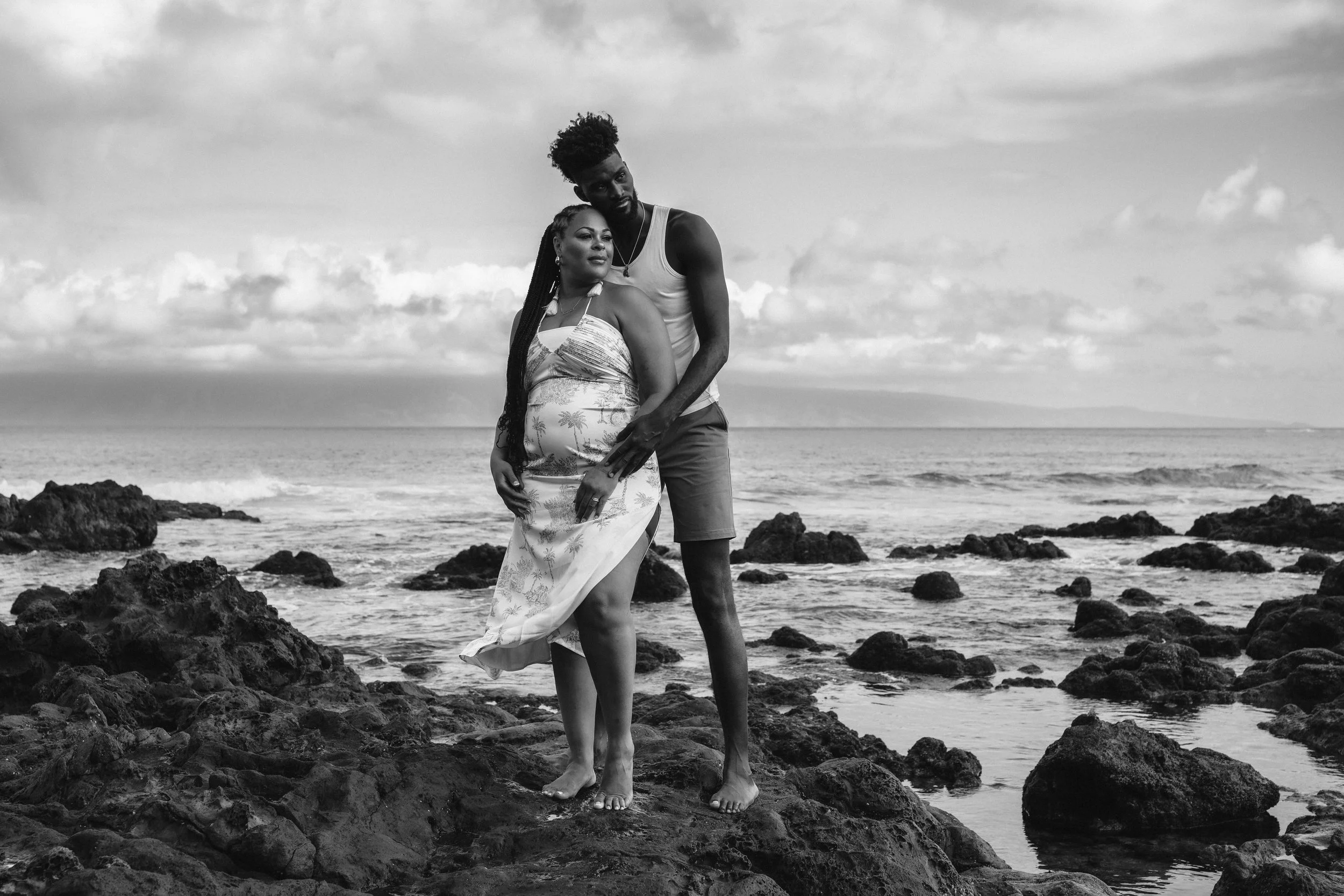Black-and-white portrait of a couple embracing on a rocky shoreline.
