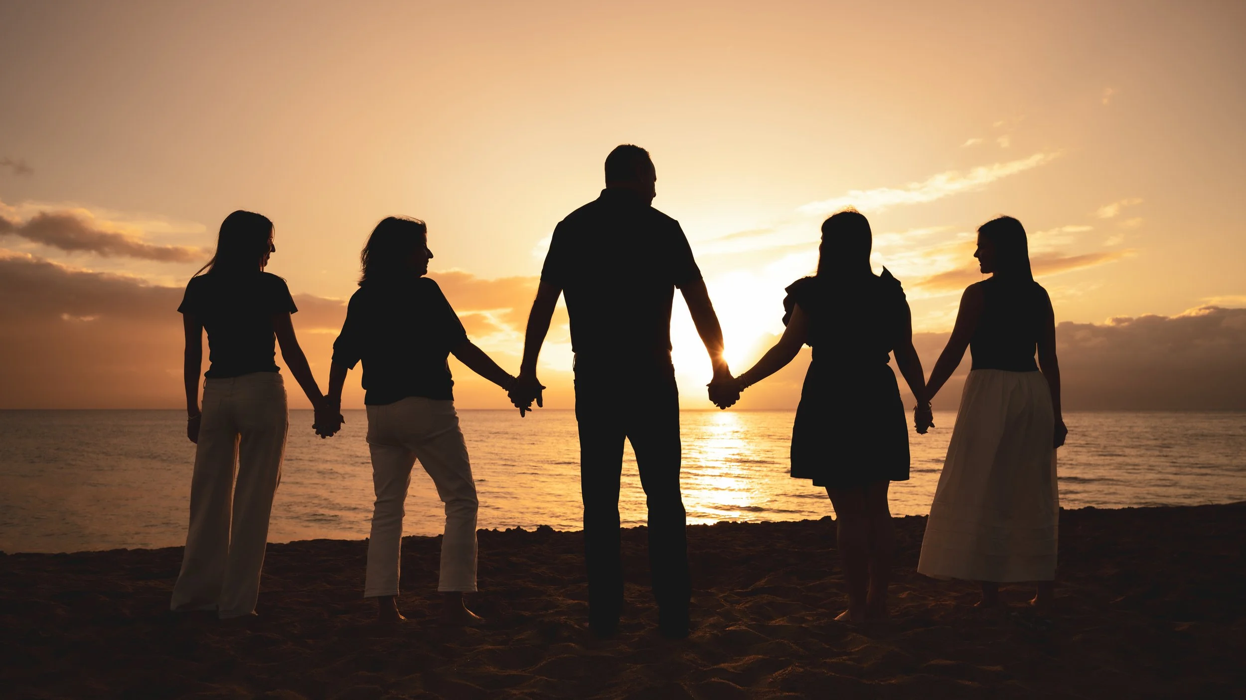 Family of five holding hands on a Maui beach, silhouetted against a golden sunset over the ocean.