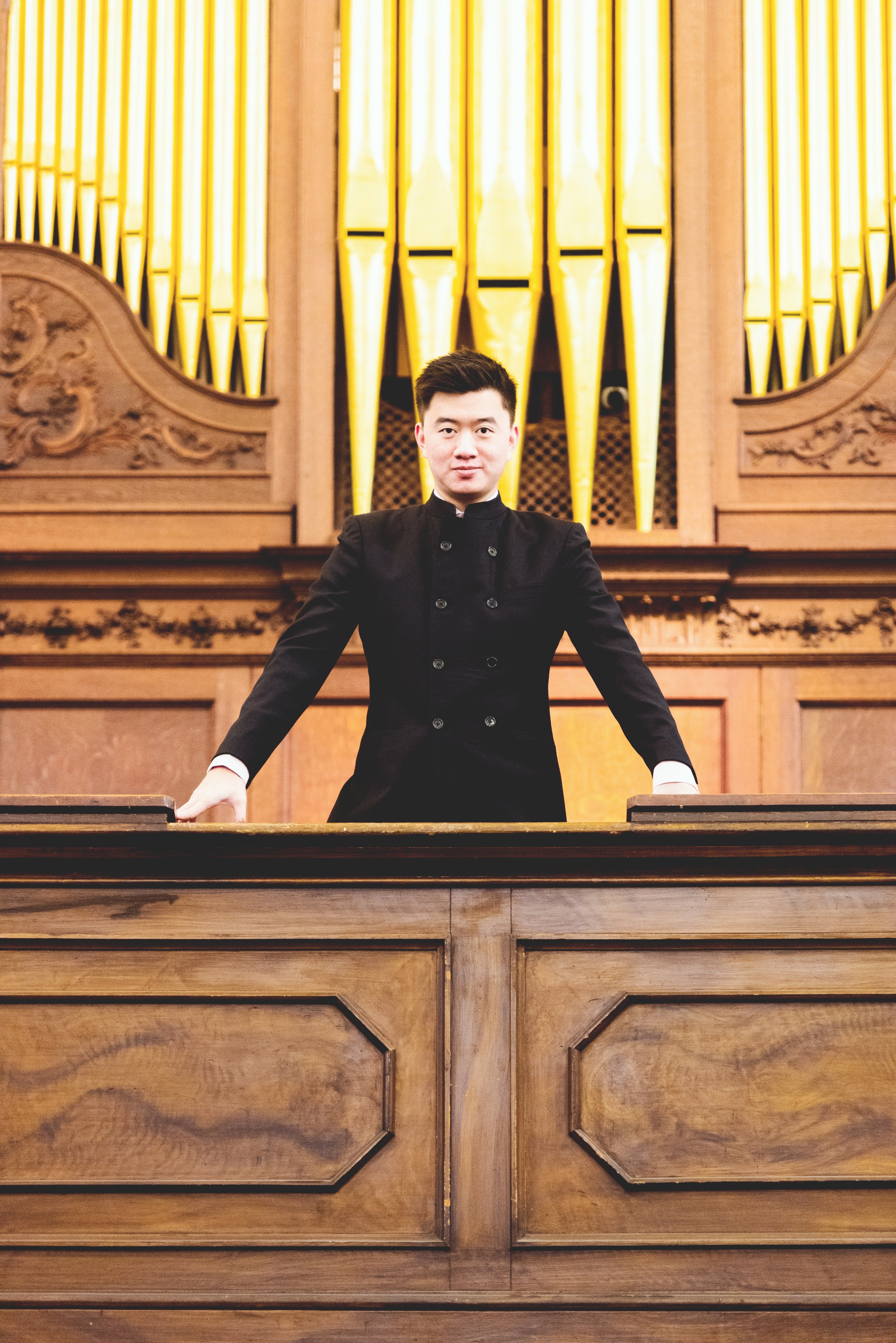 A young man in a black formal jacket standing at a church's pipe organ with ornate wooden panels behind him.