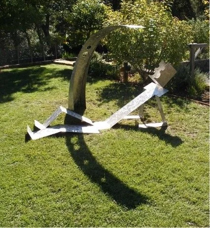 Outdoor metal sculpture of an abstract human figure beneath a curved wooden arch in a sunny grassy garden