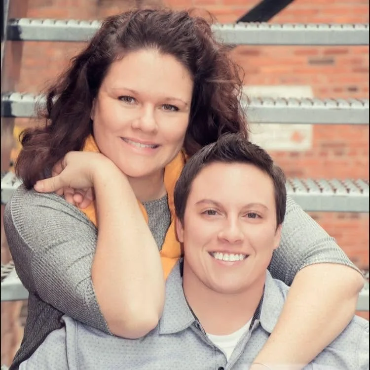 A woman with curly brown hair and a man with short dark hair sitting close together outdoors, smiling at the camera, with a brick wall and metal stairs in the background.