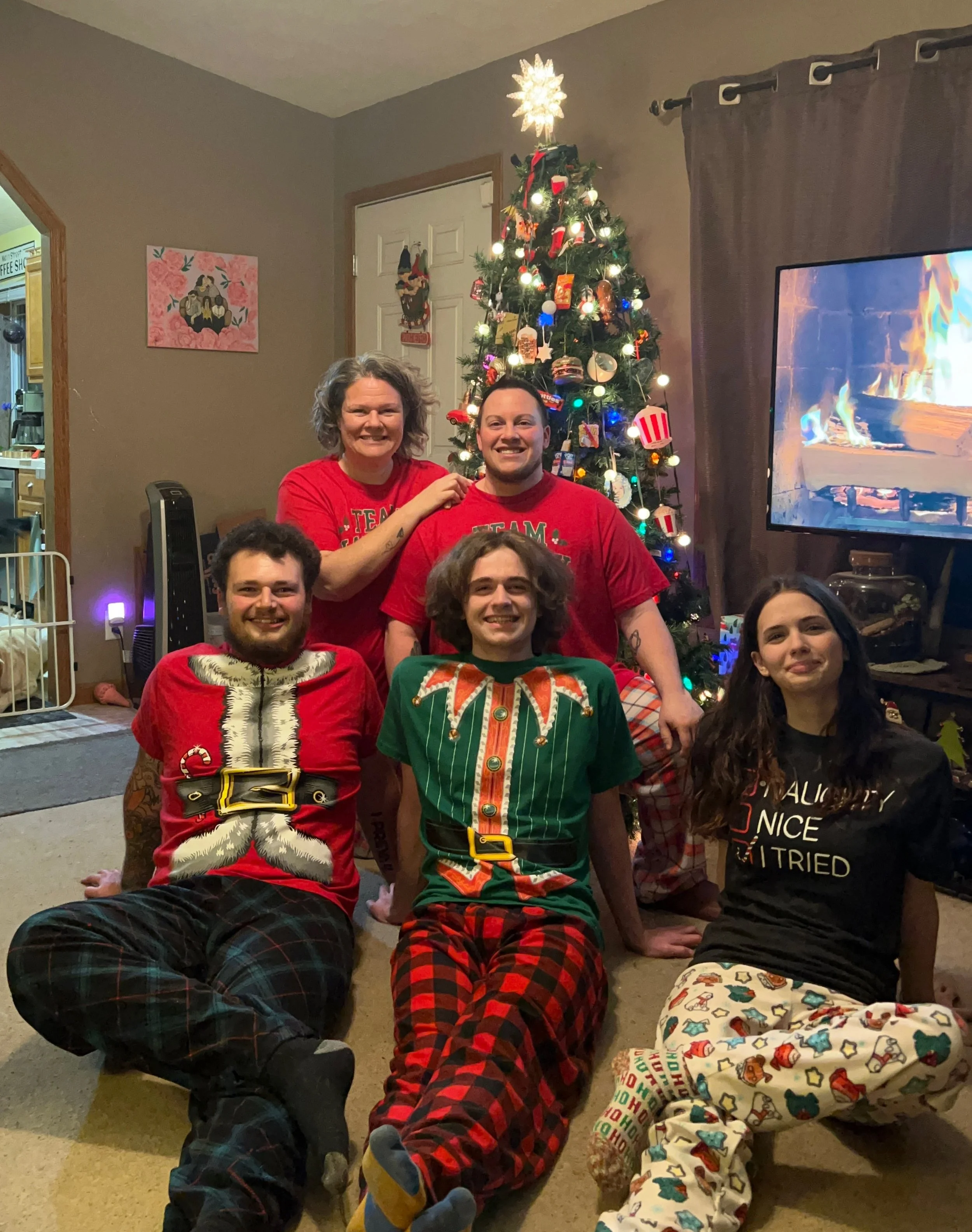A family of five celebrating Christmas in front of a decorated Christmas tree with stockings and ornaments. They are wearing festive pajamas and smiling.