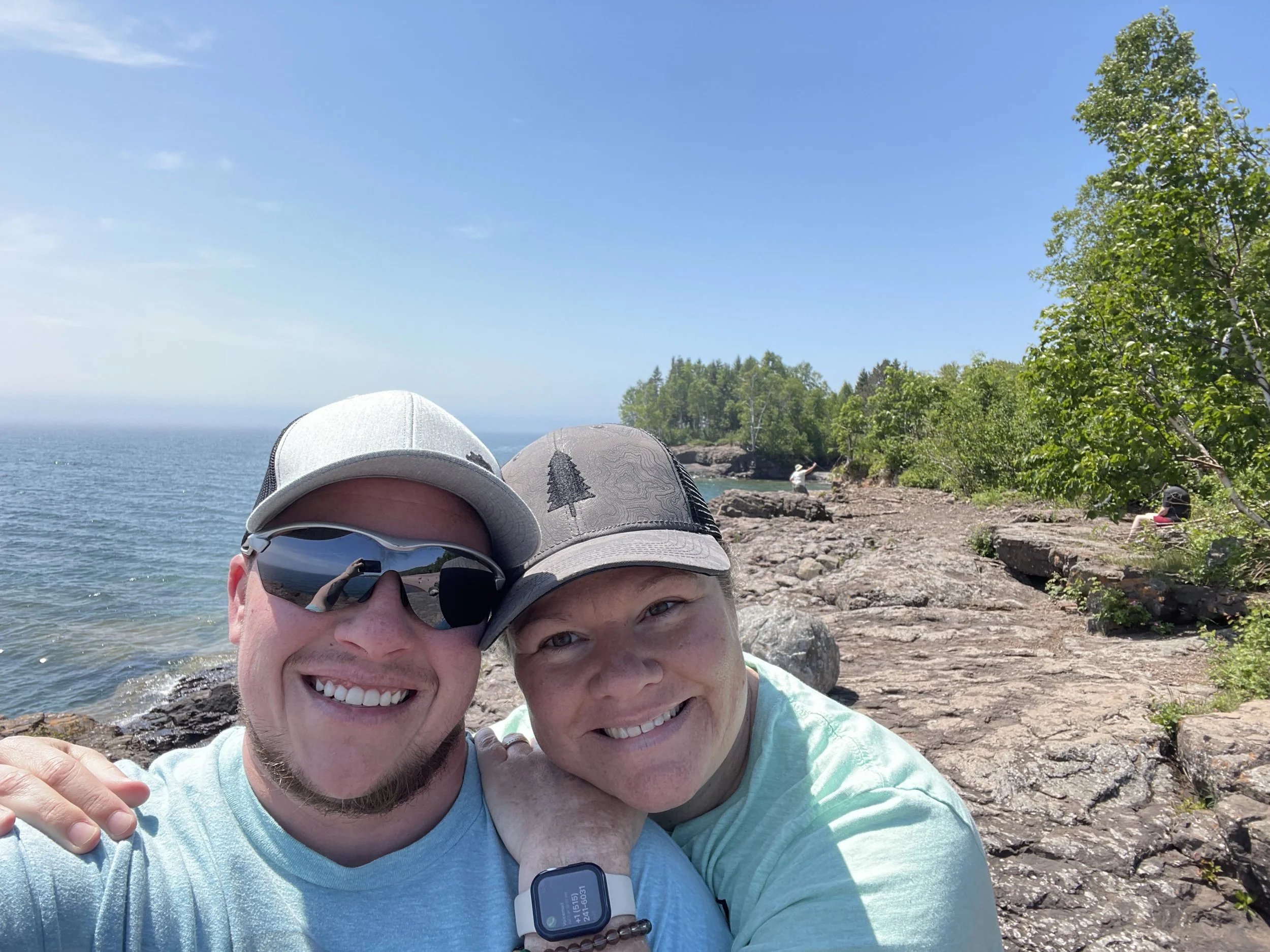 A smiling couple taking a selfie outdoors near a lake on a sunny day, with trees and rocky shoreline visible in the background. Both are wearing hats and casual clothing.