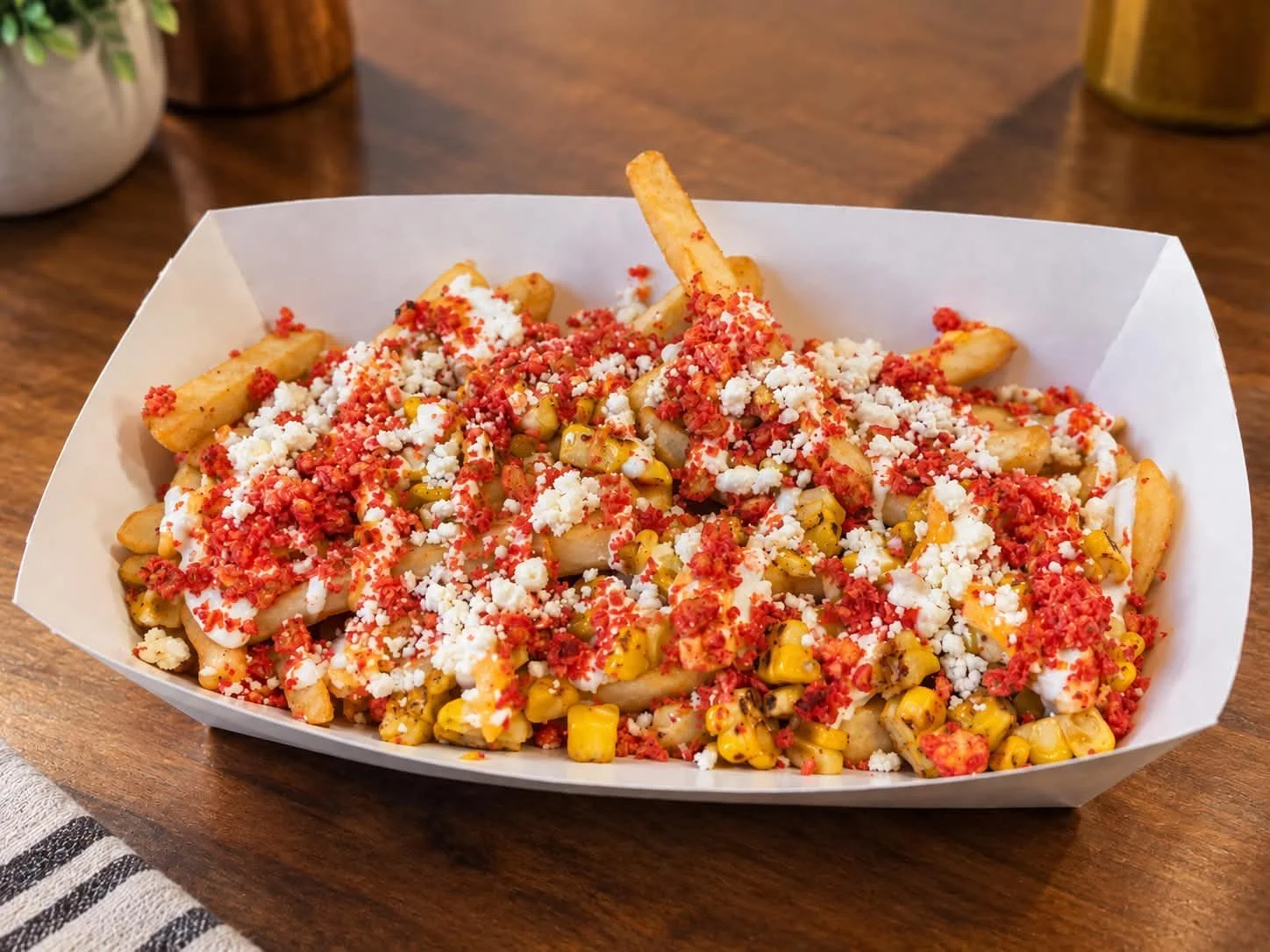 Dish of loaded French fries topped with cheese, salsa, and crumbled cheese in a paper tray on a wooden table.