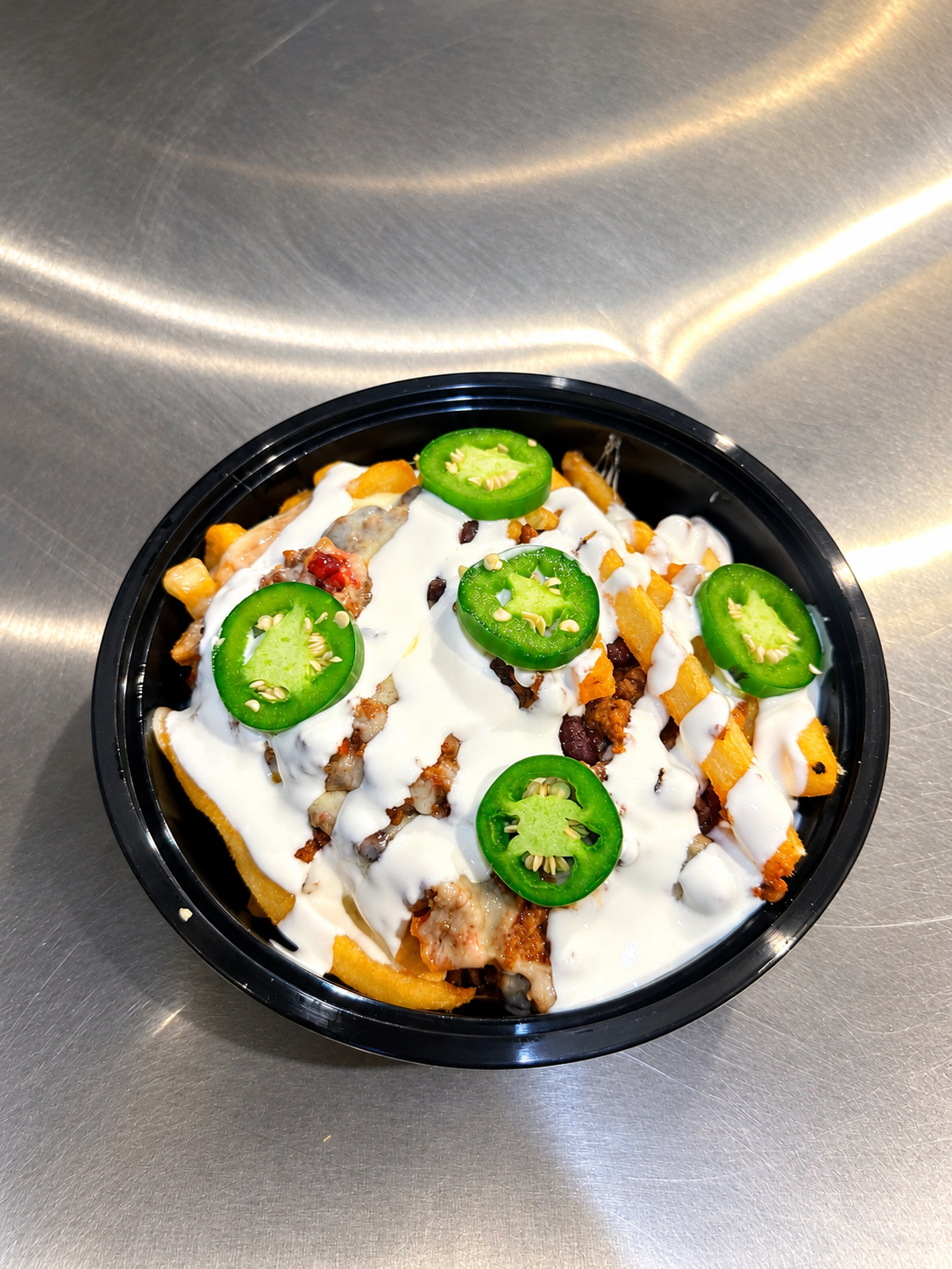 A bowl of loaded nachos topped with green jalapeño slices, drizzled with white sauce, and garnished with chopped ingredients on a stainless steel surface.