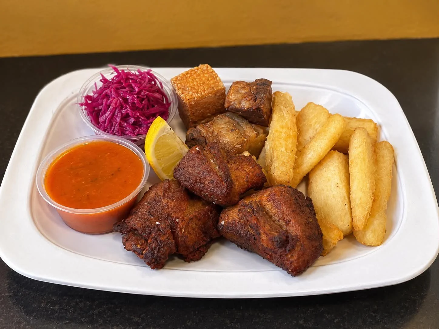 Plate of fried chicken, French fries, plantains, pickled red cabbage, a lemon wedge, tamarind candy, and hot sauce.