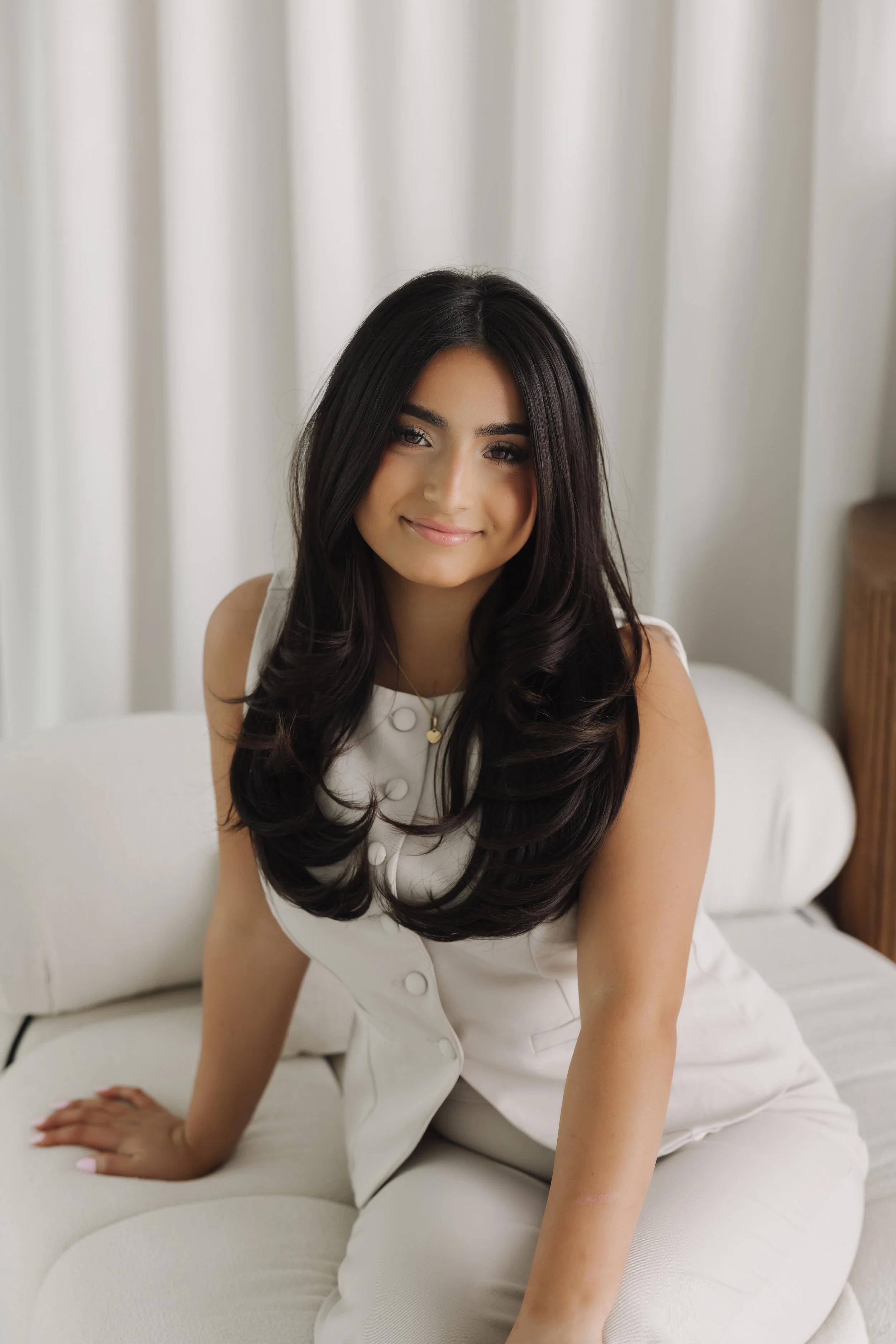 Smiling woman with long dark hair sitting on a white bed in a bright room