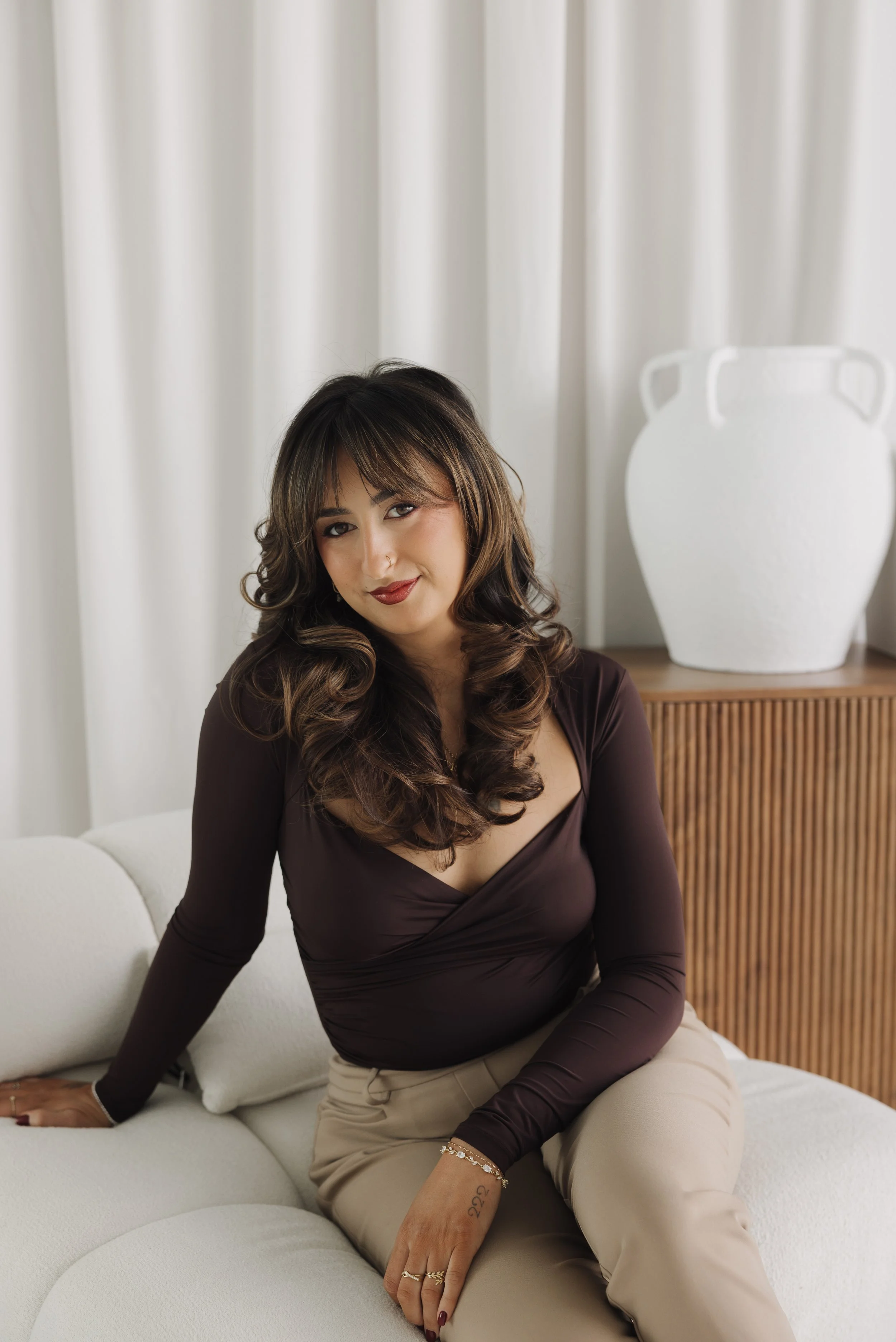Woman with curly brown hair sitting on a white sofa in a dark long-sleeve top and beige pants